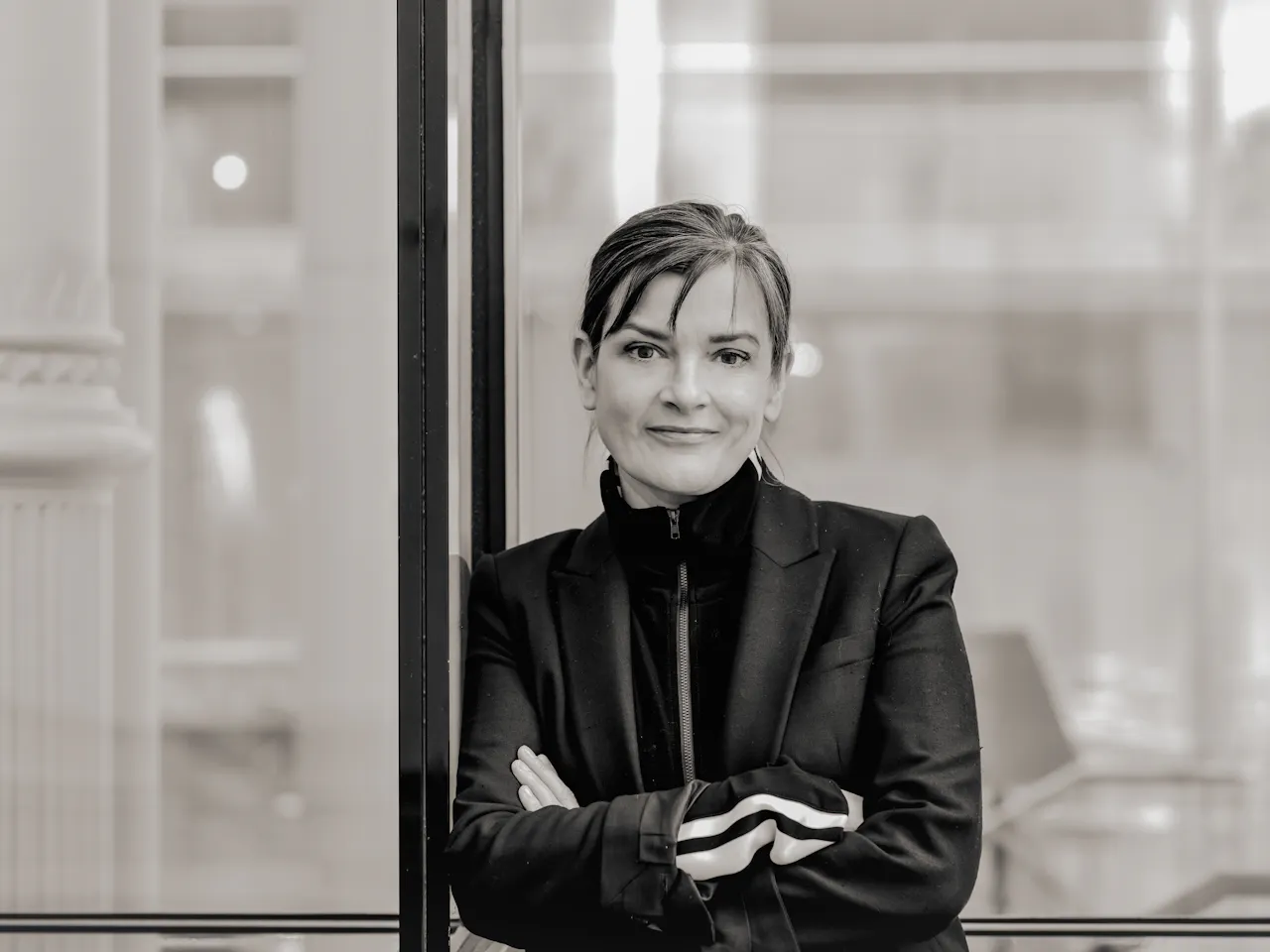 A black and white headshot of a person wearing a black jacket and black high necked jumper. It is a headshot of the Associate Director of The Royal Opera, Netia Jones.
