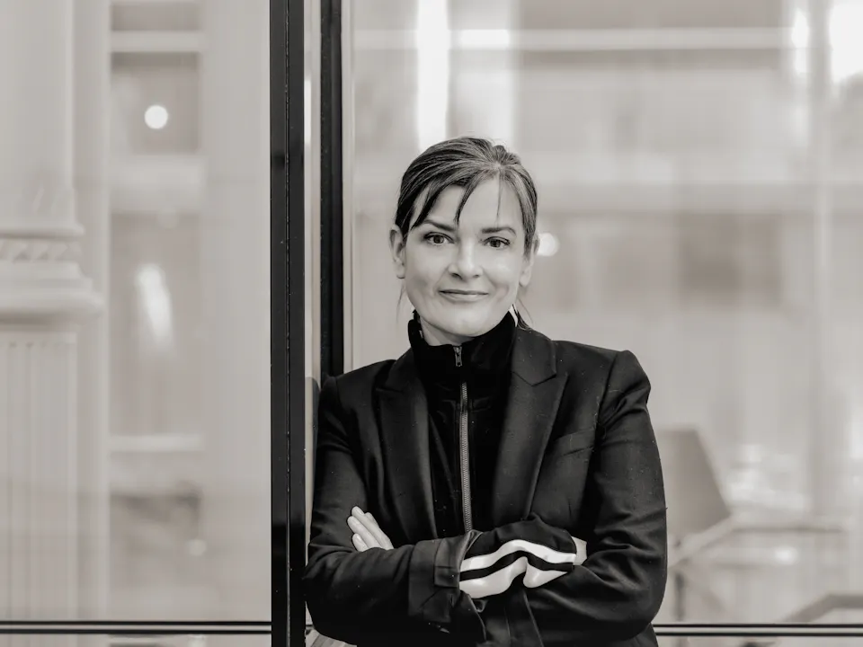 A black and white headshot of a person wearing a black jacket and black high necked jumper. It is a headshot of the Associate Director of The Royal Opera, Netia Jones.