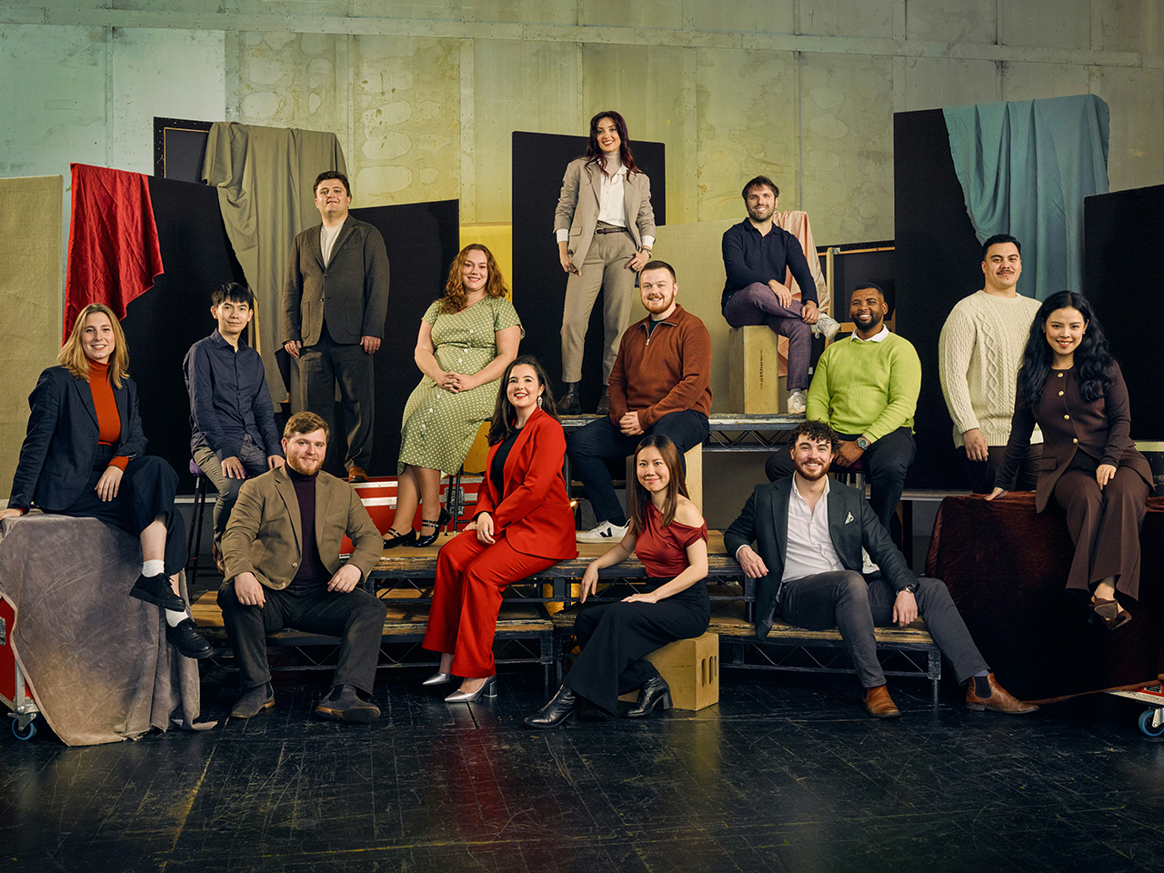A group of 13 people pose in colourful formal clothing against a textured backdrop with fabric drapes. They are seated on various platforms. 