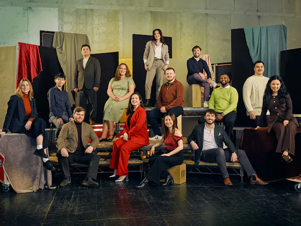 A group of 13 people pose in colourful formal clothing against a textured backdrop with fabric drapes. They are seated on various platforms.