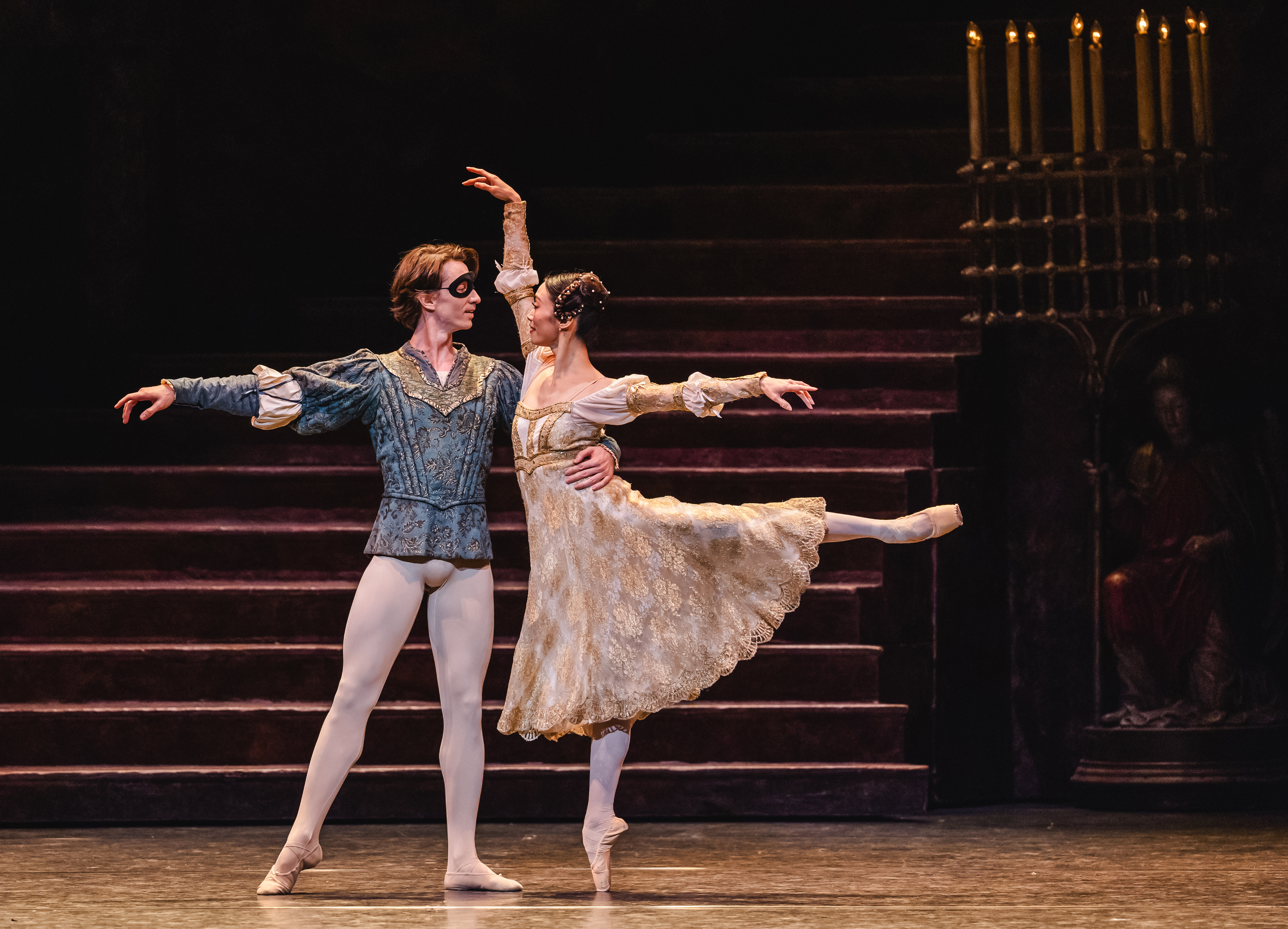 Vadim Muntagirov plays Romeo and Fumi Kaneko plays Juliet as they perform a graceful pas de deux on stage. Romeo, wearing a blue costume and a black eye mask, supports Fumi, who is dressed in a flowing cream-colored dress and balancing en pointe with one leg extended behind her. The background features a dimly lit staircase and a candelabra, creating a classical atmosphere.