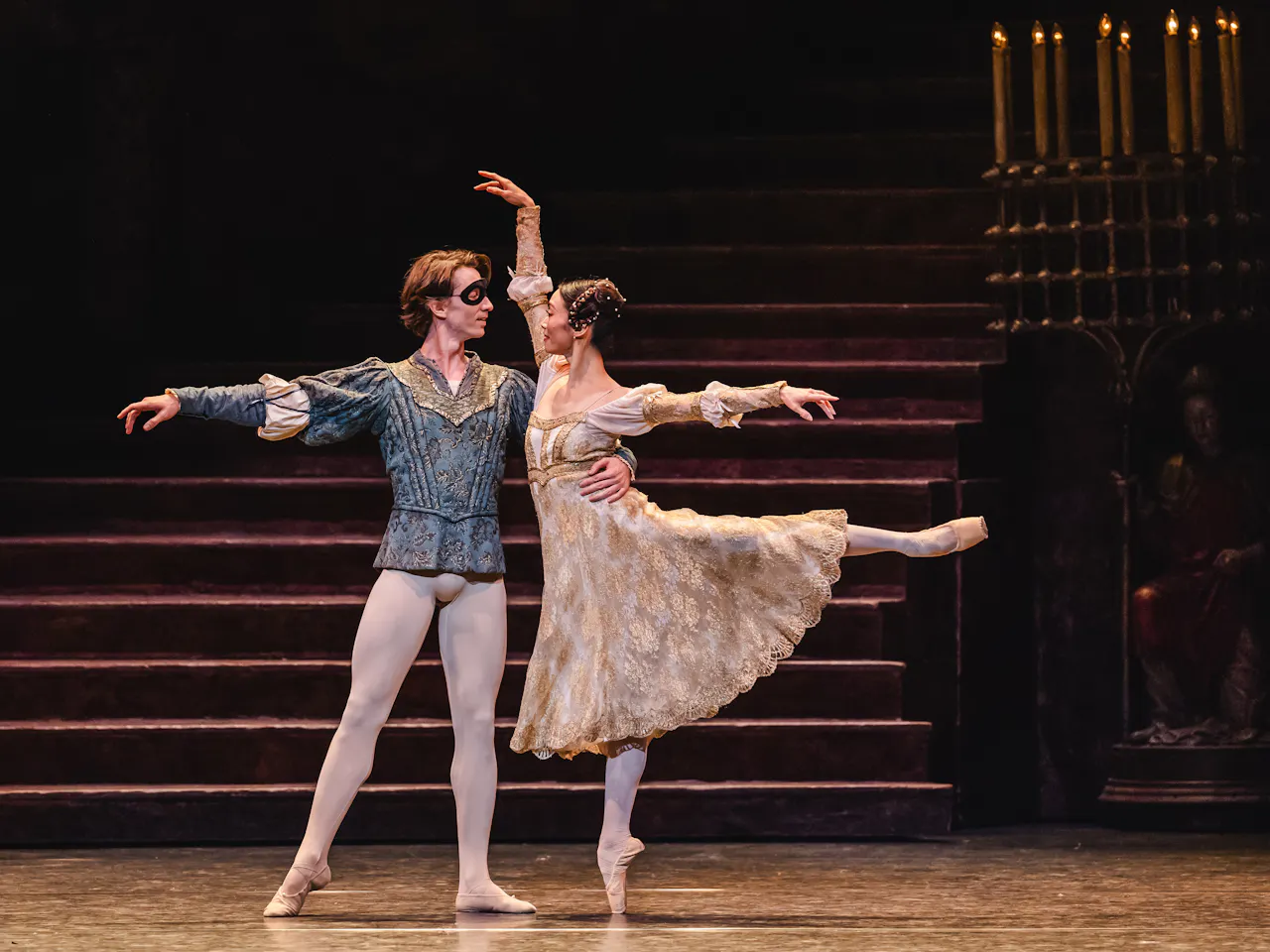 Vadim Muntagirov plays Romeo and Fumi Kaneko plays Juliet as they perform a graceful pas de deux on stage. Romeo, wearing a blue costume and a black eye mask, supports Fumi, who is dressed in a flowing cream-colored dress and balancing en pointe with one leg extended behind her. The background features a dimly lit staircase and a candelabra, creating a classical atmosphere.