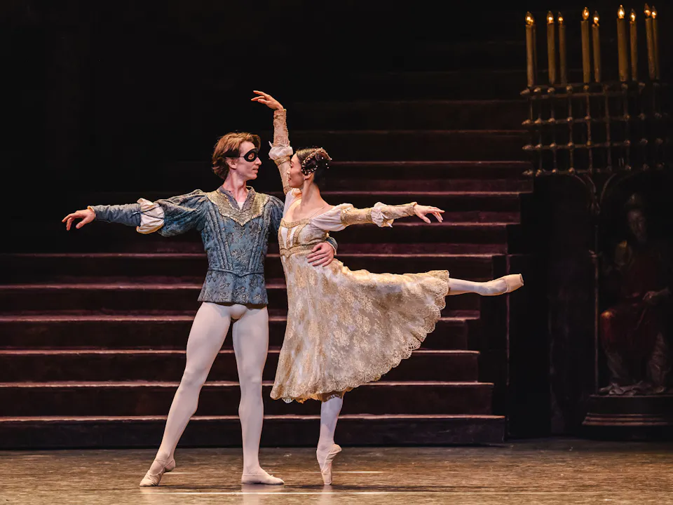 Vadim Muntagirov plays Romeo and Fumi Kaneko plays Juliet as they perform a graceful pas de deux on stage. Romeo, wearing a blue costume and a black eye mask, supports Fumi, who is dressed in a flowing cream-colored dress and balancing en pointe with one leg extended behind her. The background features a dimly lit staircase and a candelabra, creating a classical atmosphere.
