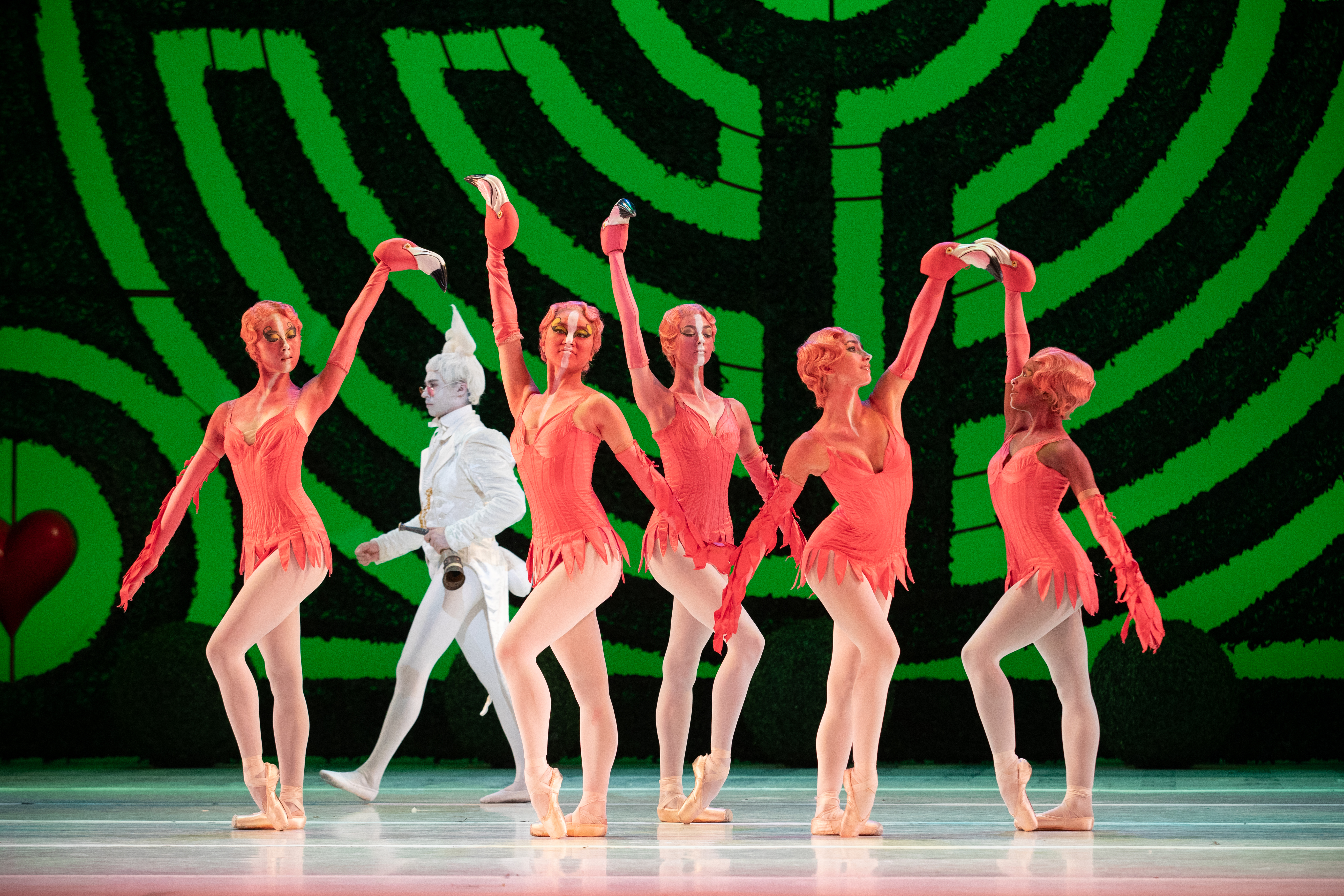 A group of dancers dressed as flamingos stand on stage with arms raised. Behind them is a backdrop with a large green maze. (2 of 6)