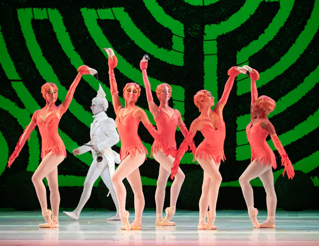A group of dancers dressed as flamingos stand on stage with arms raised. Behind them is a backdrop with a large green maze. (2 of 6)