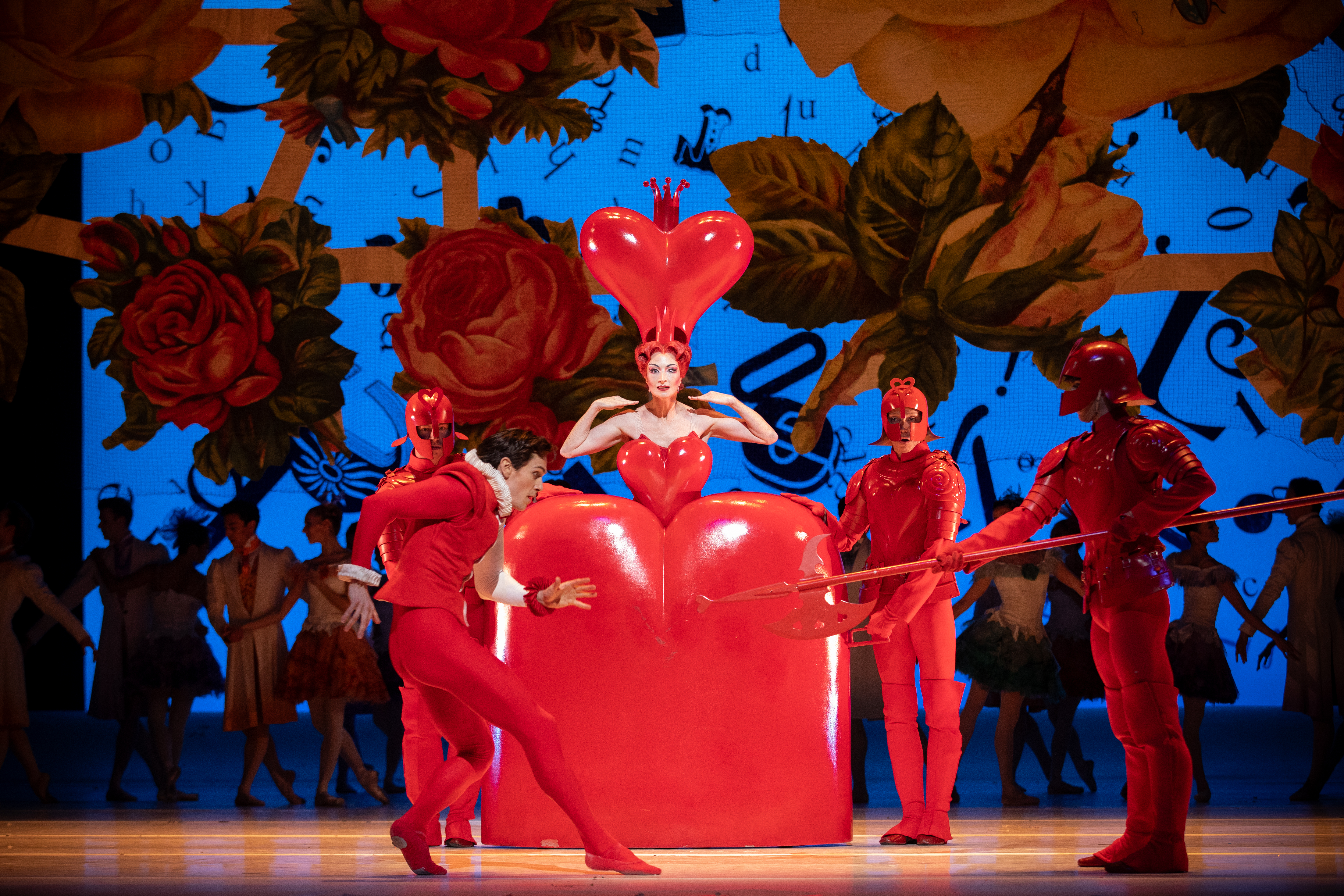 The Queen of Hearts stands in the centre of the stage, she wears a large, heart-shaped red dress. Three guards in red uniform stand around her. In front a dancer dressed as the Knave of Hearts creeps forward. (3 of 6)