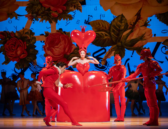 The Queen of Hearts stands in the centre of the stage, she wears a large, heart-shaped red dress. Three guards in red uniform stand around her. In front a dancer dressed as the Knave of Hearts creeps forward. (3 of 6)