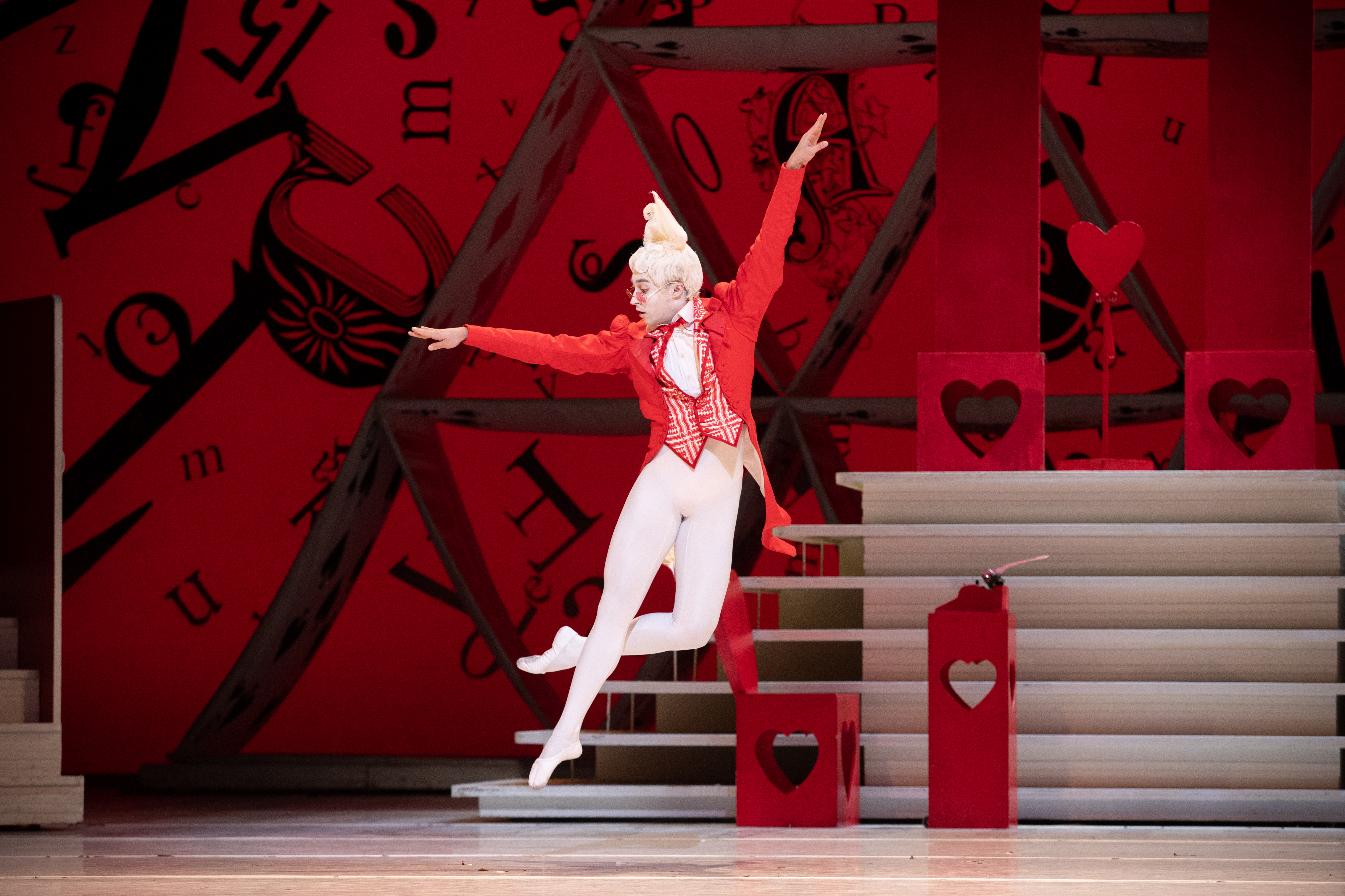 A dancer with white rabbit ears, a red jacket and white tights jumps on a stage. His arms are outstretched and behind him is a large red backdrop. (1 of 6)