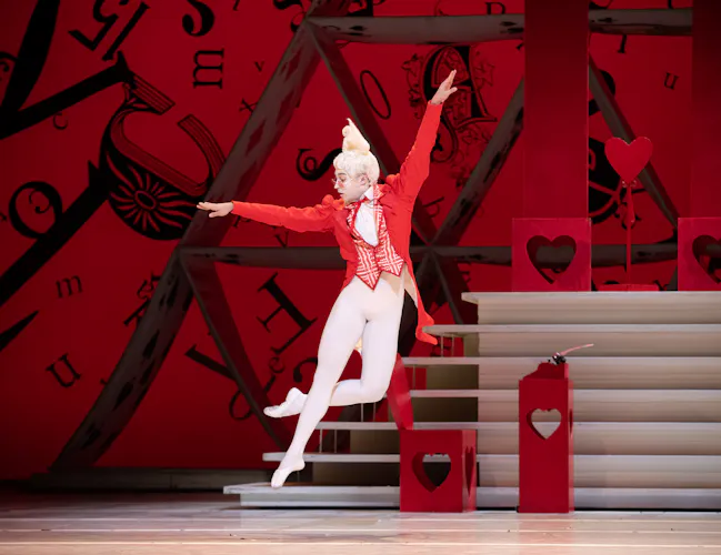 A dancer with white rabbit ears, a red jacket and white tights jumps on a stage. His arms are outstretched and behind him is a large red backdrop. (1 of 6)