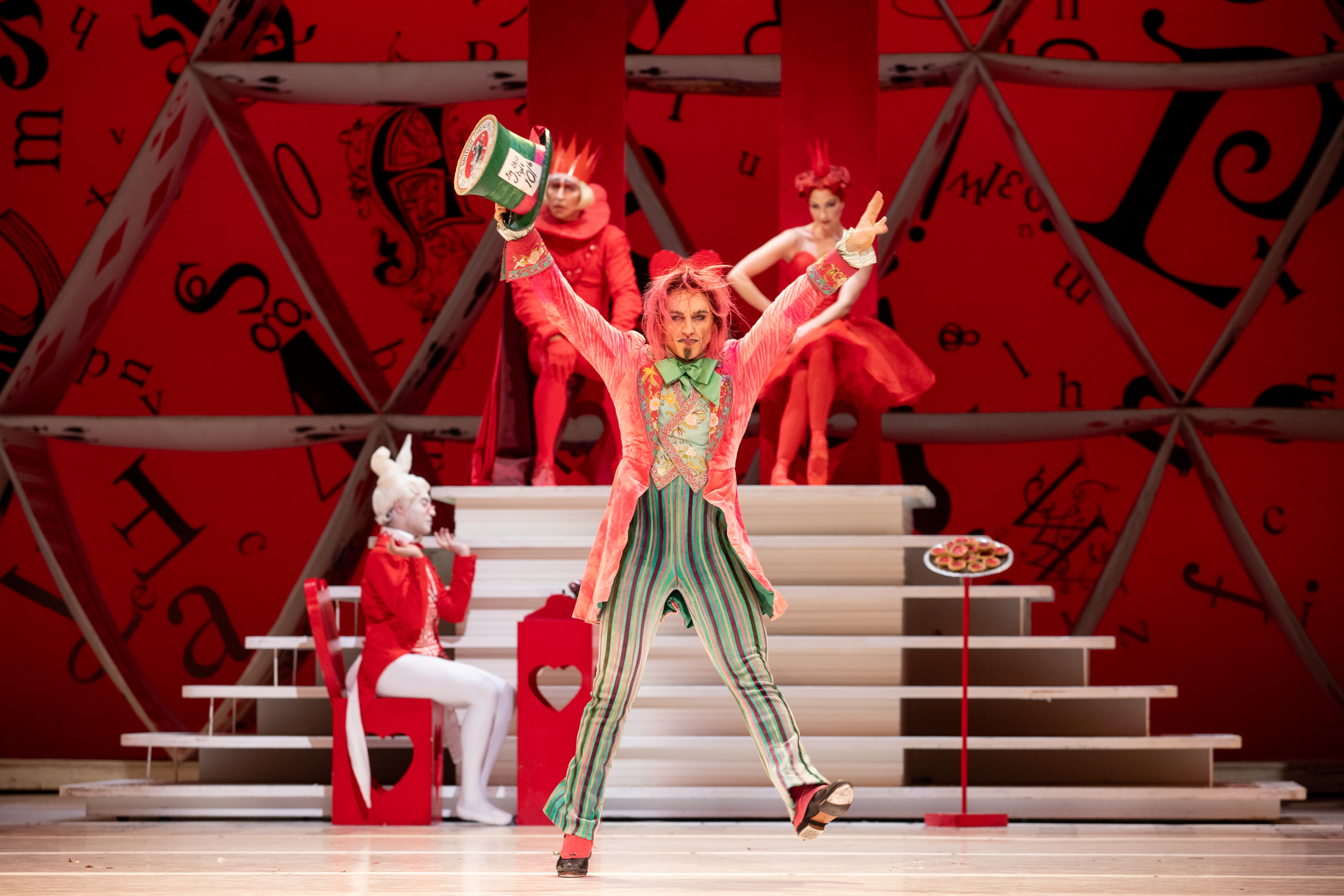Steven McRae as The Mad Hatter stands in the centre of the stage, he wears a long pink coat, striped green trousers and waistcoat and holds a large green top hat in his outstretched hand. Behind him is a podium upon which the king and queen of hearts sit. In front o the podium the white rabbit is sat at a table taking notes. (6 of 6)