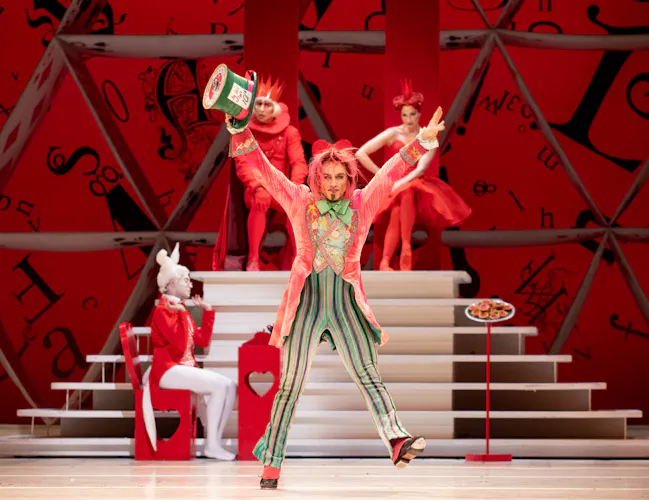 Steven McRae as The Mad Hatter stands in the centre of the stage, he wears a long pink coat, striped green trousers and waistcoat and holds a large green top hat in his outstretched hand. Behind him is a podium upon which the king and queen of hearts sit. In front o the podium the white rabbit is sat at a table taking notes. (6 of 6)
