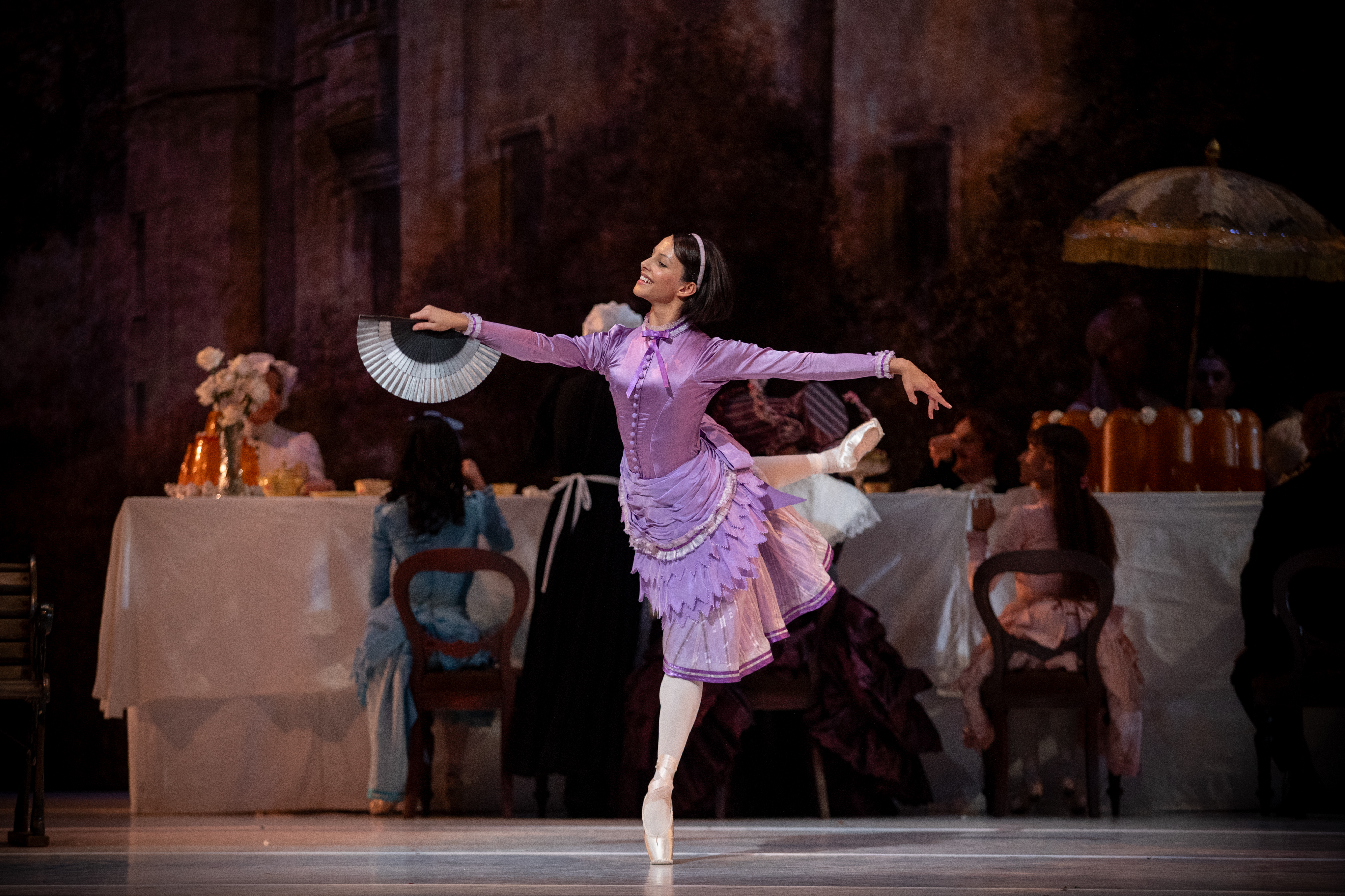 Francesca Hayward as Alice stands en pointe on one leg on stage. She is wearing a purple dress. Behind her a long table is set up for a picnic. (4 of 6)
