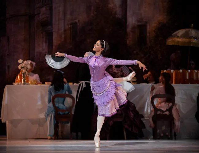 Francesca Hayward as Alice stands en pointe on one leg on stage. She is wearing a purple dress. Behind her a long table is set up for a picnic. (4 of 6)