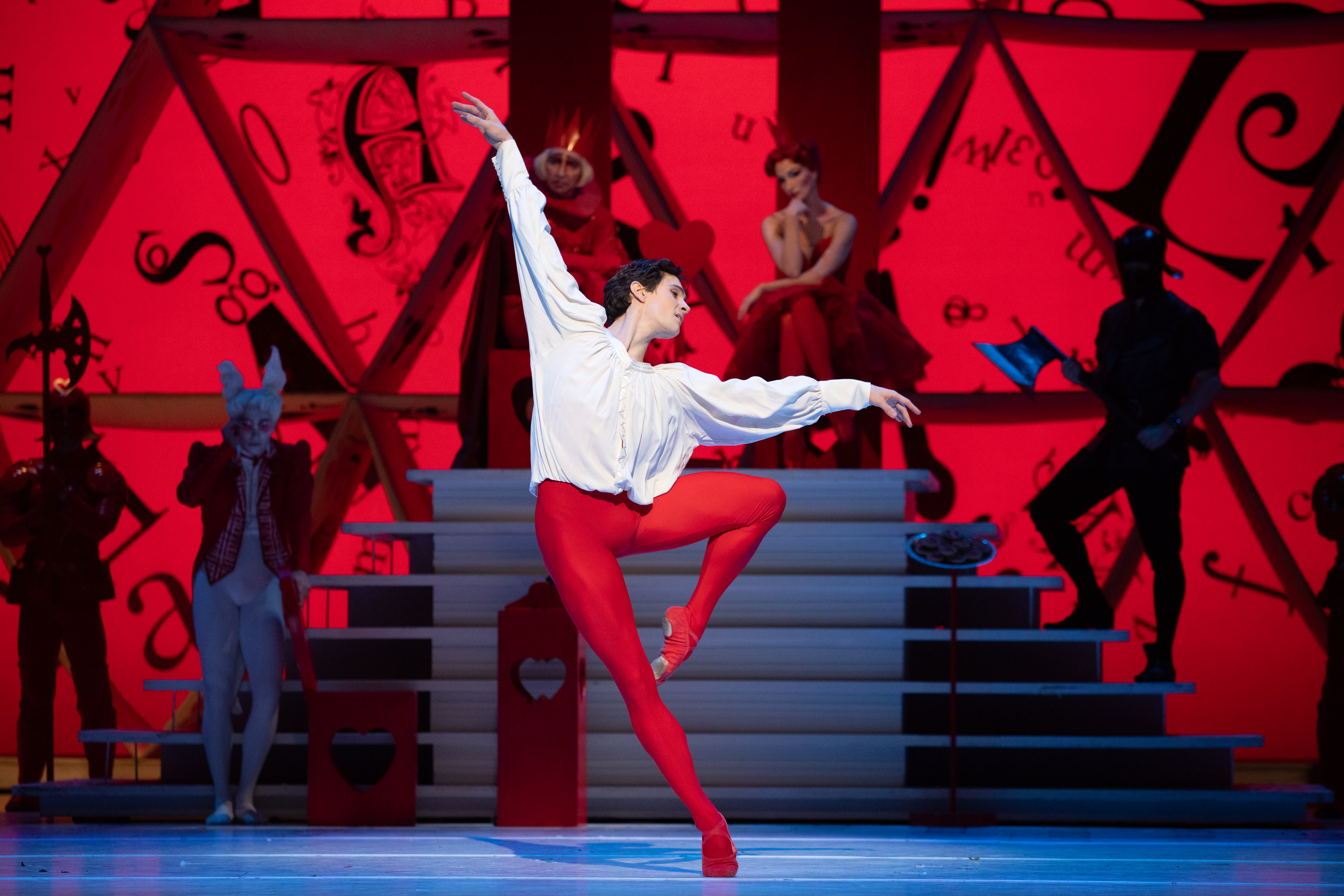 William Bracewell as The Knave of Hearts stands on one leg in the centre of the stage. He wears red tights and a white shirt. Behind him is a podium upon which the king and queen of hearts sit, surrounded by guards. (5 of 6)