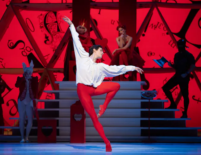 William Bracewell as The Knave of Hearts stands on one leg in the centre of the stage. He wears red tights and a white shirt. Behind him is a podium upon which the king and queen of hearts sit, surrounded by guards. (5 of 6)