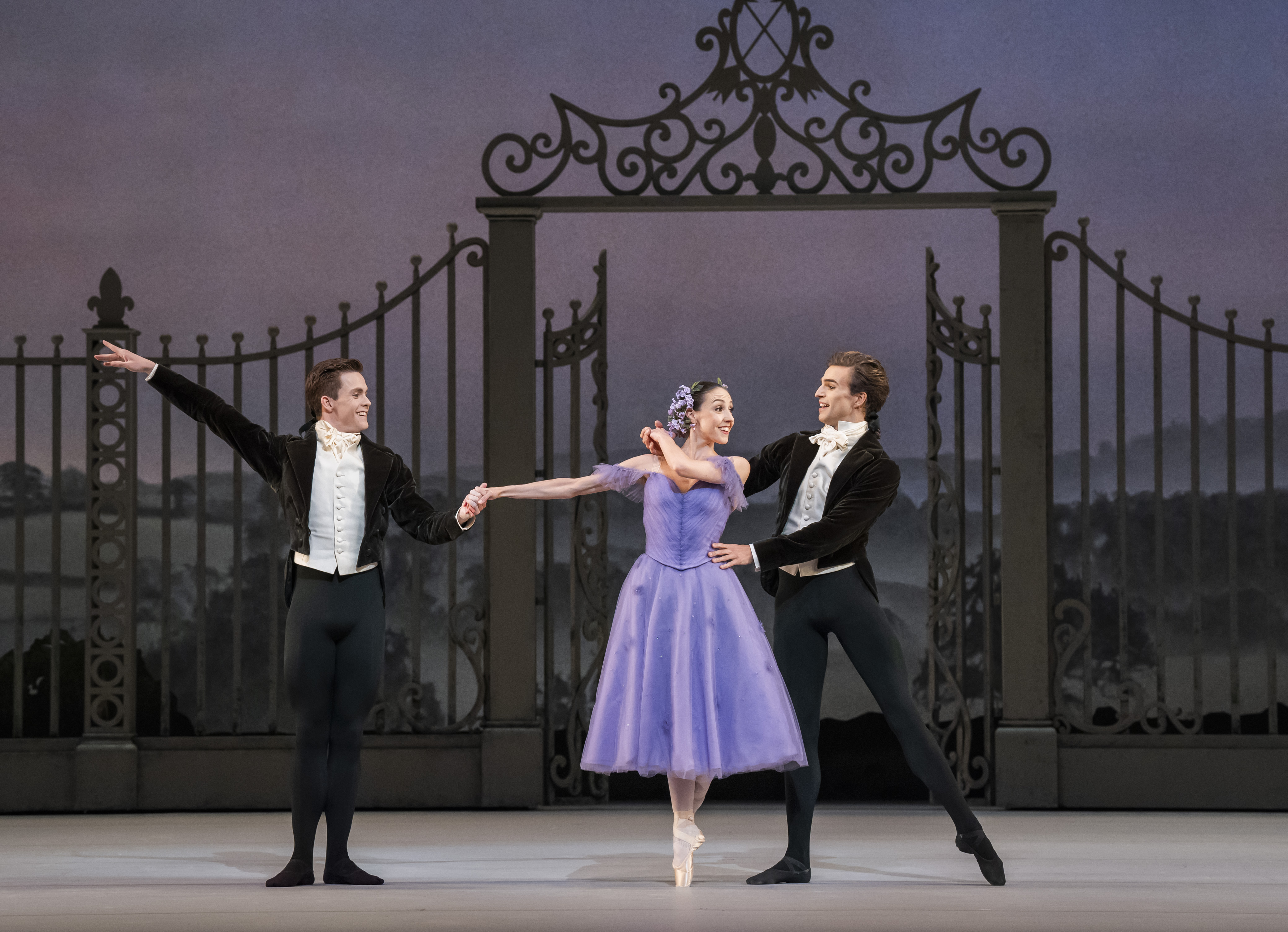 Harrison Lee, Isabella Gasparini and Leo Dixon stand in front of a large metal gate on stage. Gasparini wears a purple dress and Lee and Dixon wear black suits. Gasparini stands en pointe with her arm outstretched. (2 of 4)