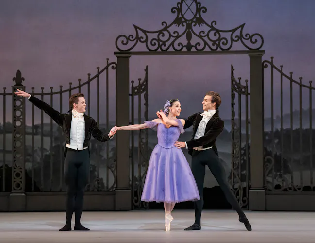 Harrison Lee, Isabella Gasparini and Leo Dixon stand in front of a large metal gate on stage. Gasparini wears a purple dress and Lee and Dixon wear black suits. Gasparini stands en pointe with her arm outstretched. (2 of 4)