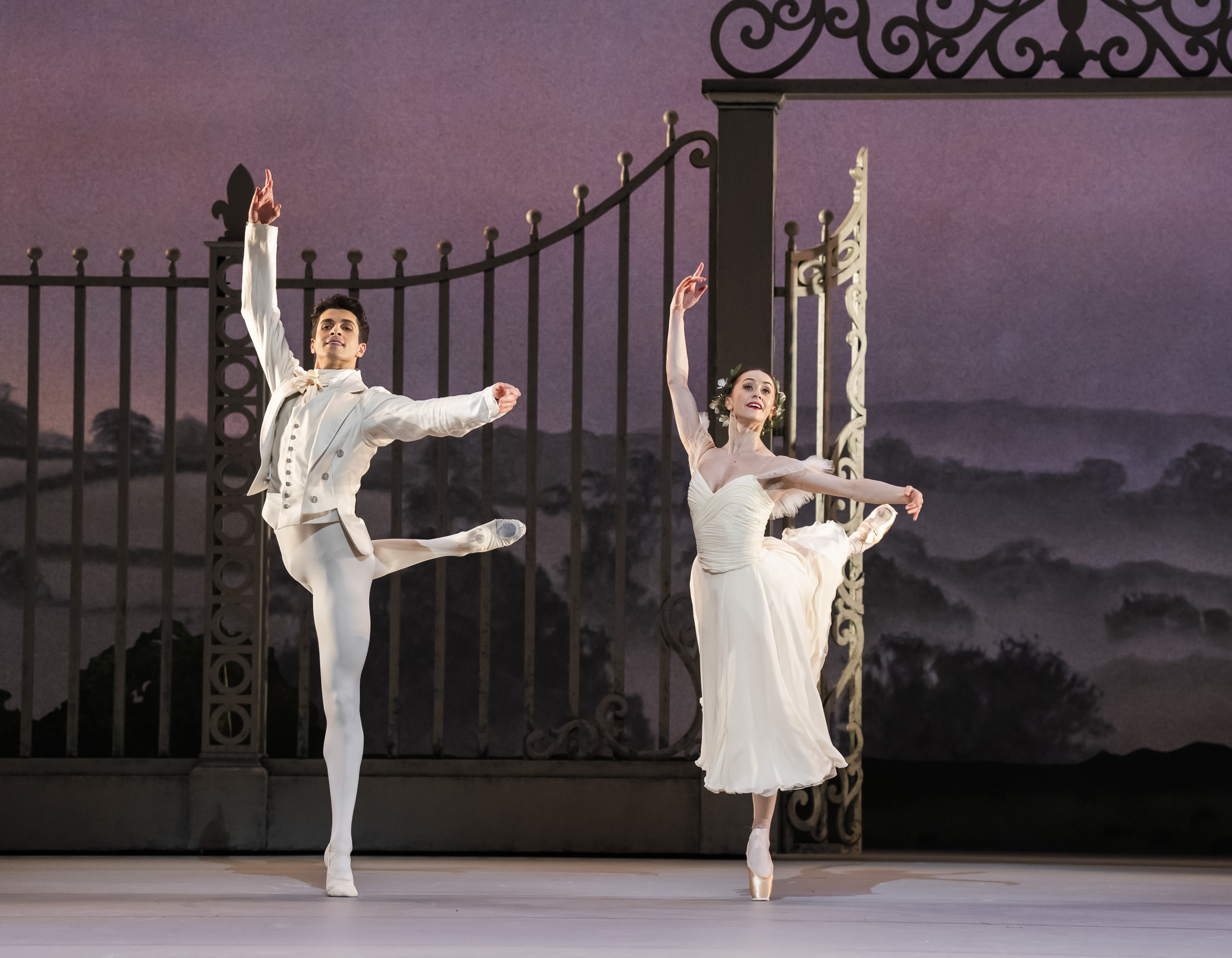 Two dancers dance in front of a large metal gate on stage. Reece Clarke stands in attitude on demi-pointe. Marianela Nunez stands in attitude en-pointe. Clarke wears a white suit and Nunez a white dress. (1 of 4)