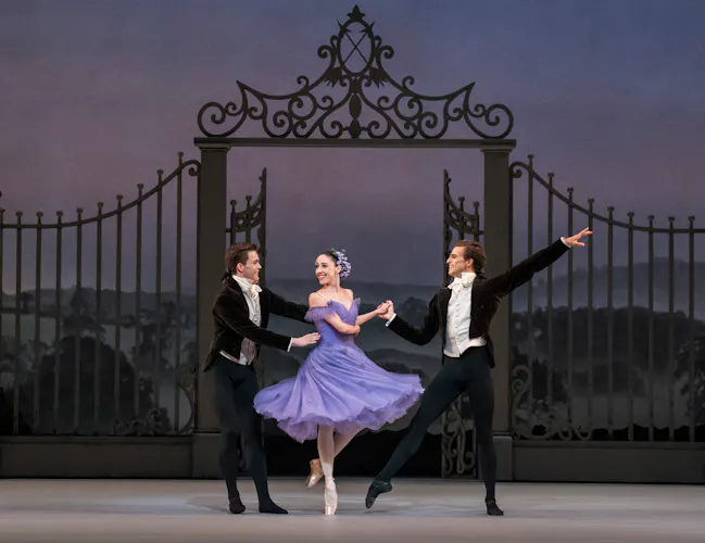Harrison Lee, Isabella Gasparini and Leo Dixon stand in front of a large metal gate on stage. Gasparini wears a purple dress and Lee and Dixon wear black suits. Gasparini stands en pointe with Lee and Dixon either side holding her. (4 of 4)