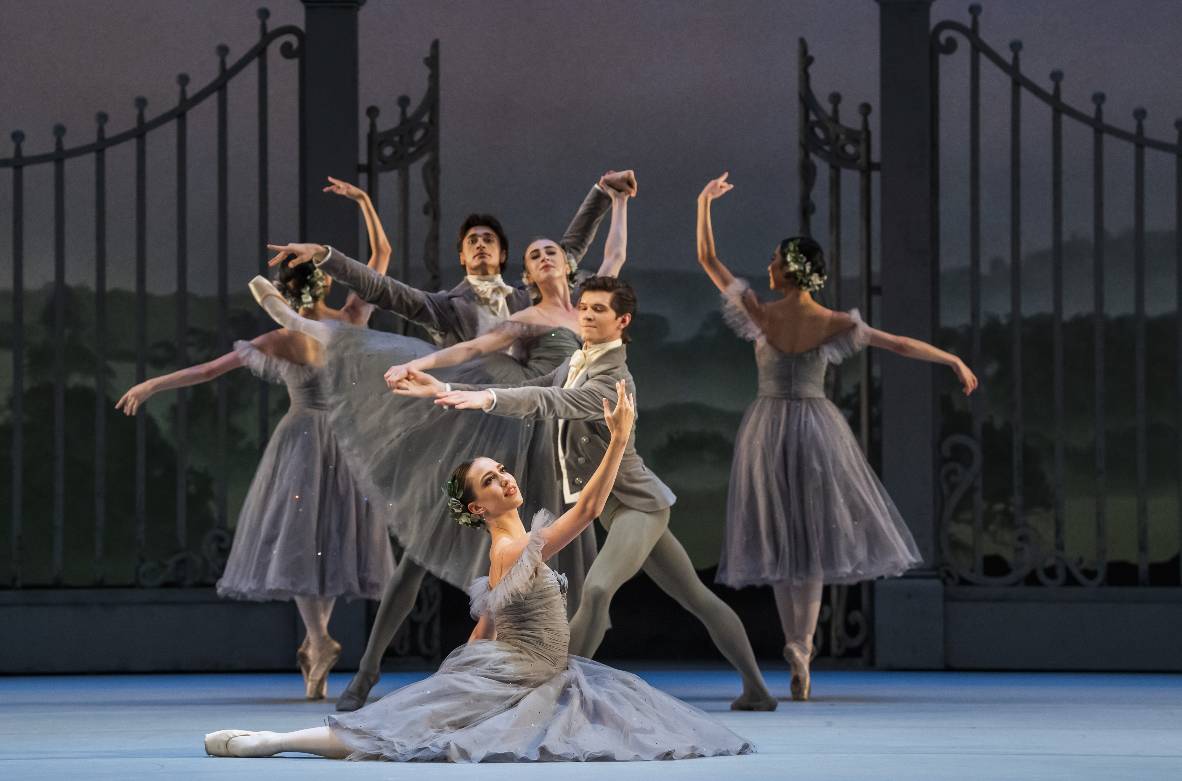 A group of dancers stand in front of a large metal gate. They hold various poses with arms outstretched. They wear grey dresses and suits. (3 of 4)