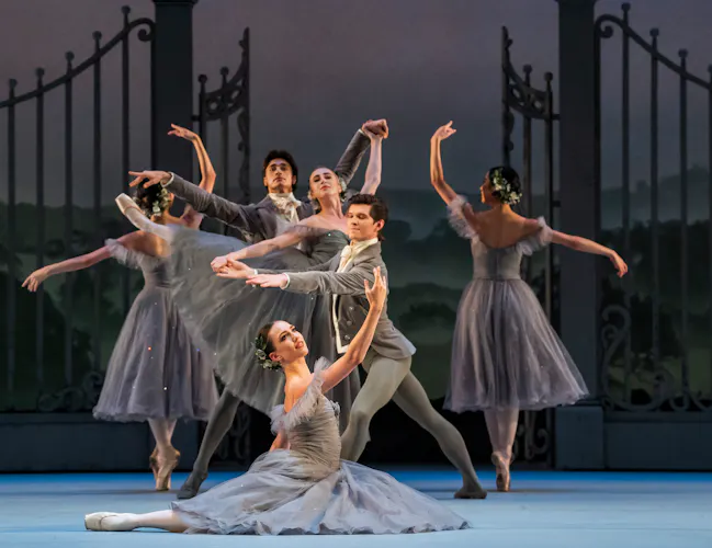 A group of dancers stand in front of a large metal gate. They hold various poses with arms outstretched. They wear grey dresses and suits. (3 of 4)