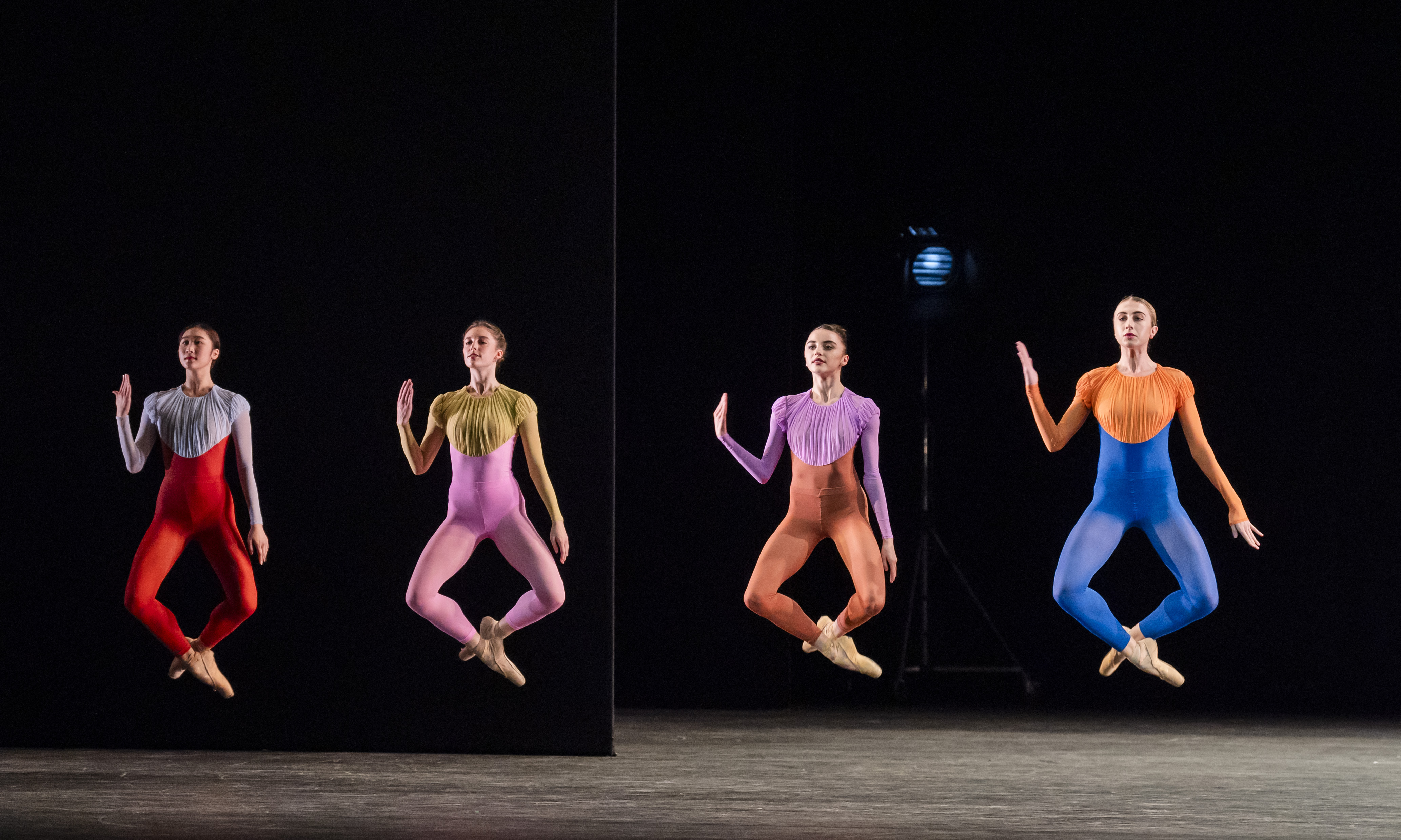 Artists of The Royal Ballet perform on a dark stage. Two dancers in colourful unitards are mid-jump. Their ankles are crossed and they all hold up their right arm. (4 of 5)