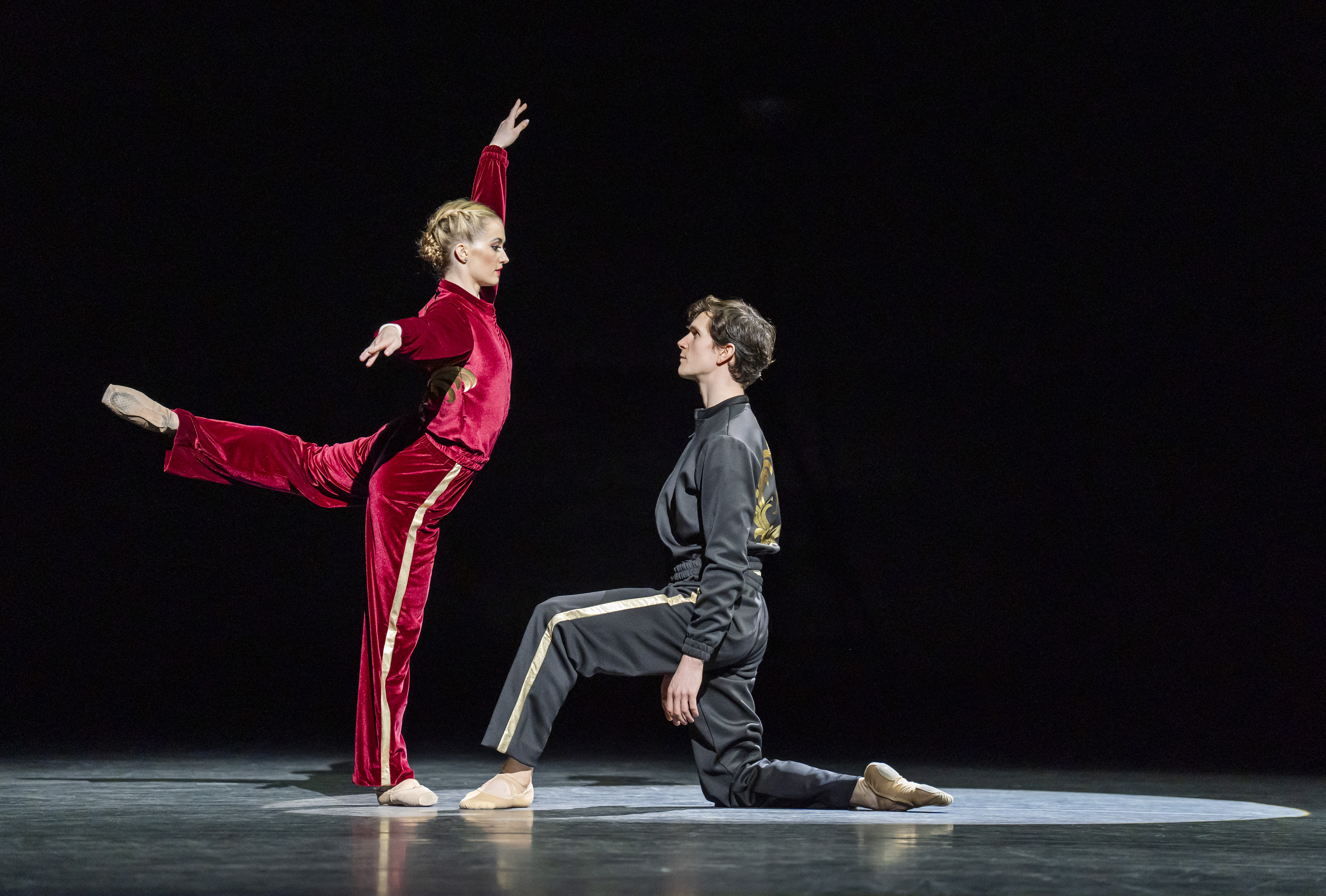 Anna Rose O'Sullivan and William Bracewell perform on a dark stage. O'Sullivan wears a red velvet tracksuit and stand on one leg, her other stretched out behind her. Bracewell wears a grey velvet tracksuit and kneels before her on one knee. (1 of 5)