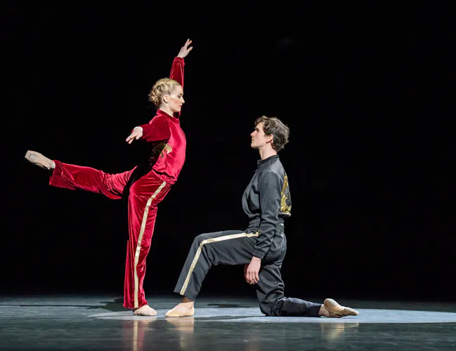 Anna Rose O'Sullivan and William Bracewell perform on a dark stage. O'Sullivan wears a red velvet tracksuit and stand on one leg, her other stretched out behind her. Bracewell wears a grey velvet tracksuit and kneels before her on one knee. (1 of 5)