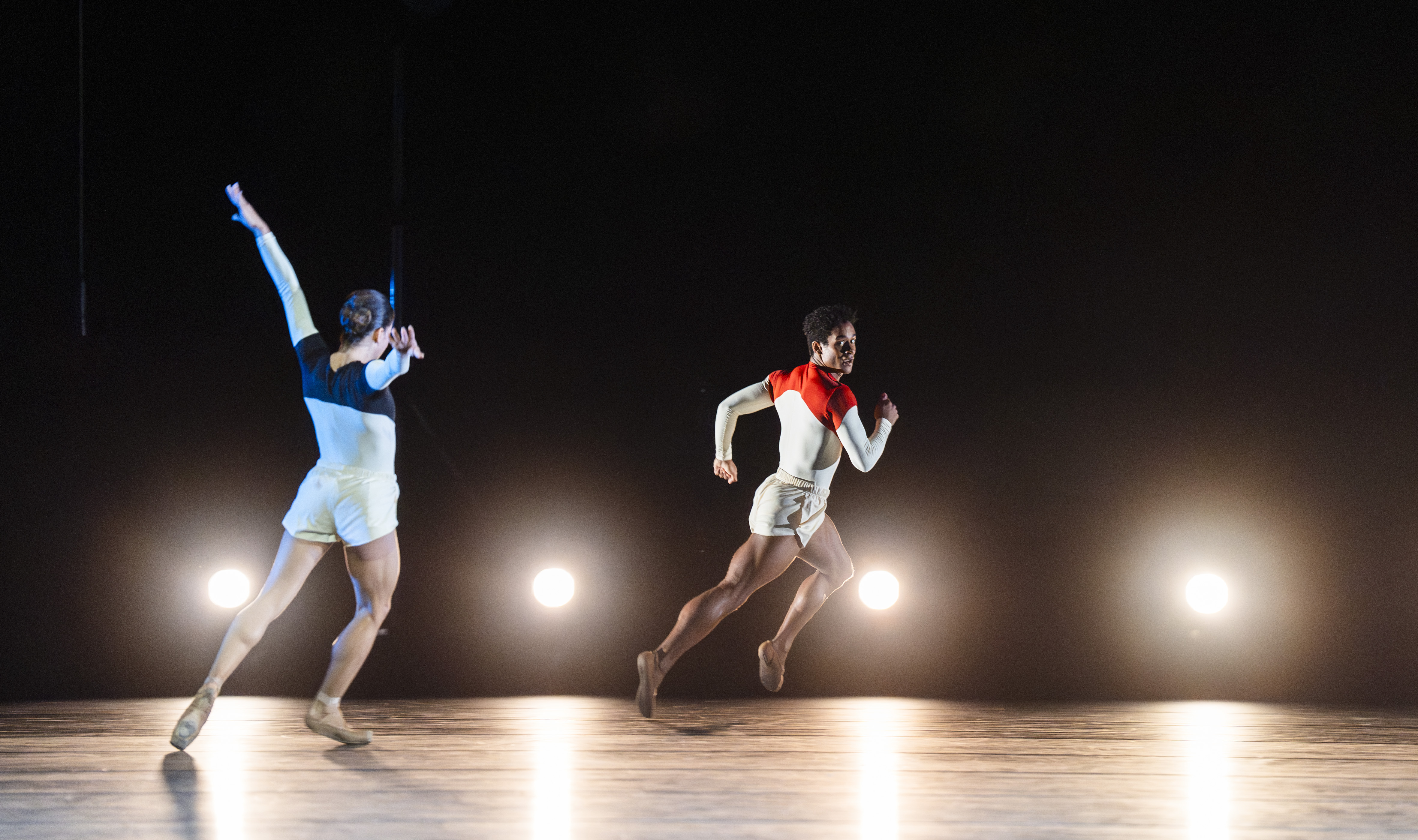 Mayara Magri and Marcelino Sambé perform on stage. Behind them is a row of lights on the floor. Magri wears white shorts and a blue and white leotard. She stands with arms outstretched. Sambé is running across the stage, he wears white shorts and red and white leotard. (5 of 5)
