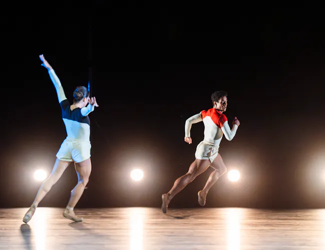 Mayara Magri and Marcelino Sambé perform on stage. Behind them is a row of lights on the floor. Magri wears white shorts and a blue and white leotard. She stands with arms outstretched. Sambé is running across the stage, he wears white shorts and red and white leotard. (5 of 5)