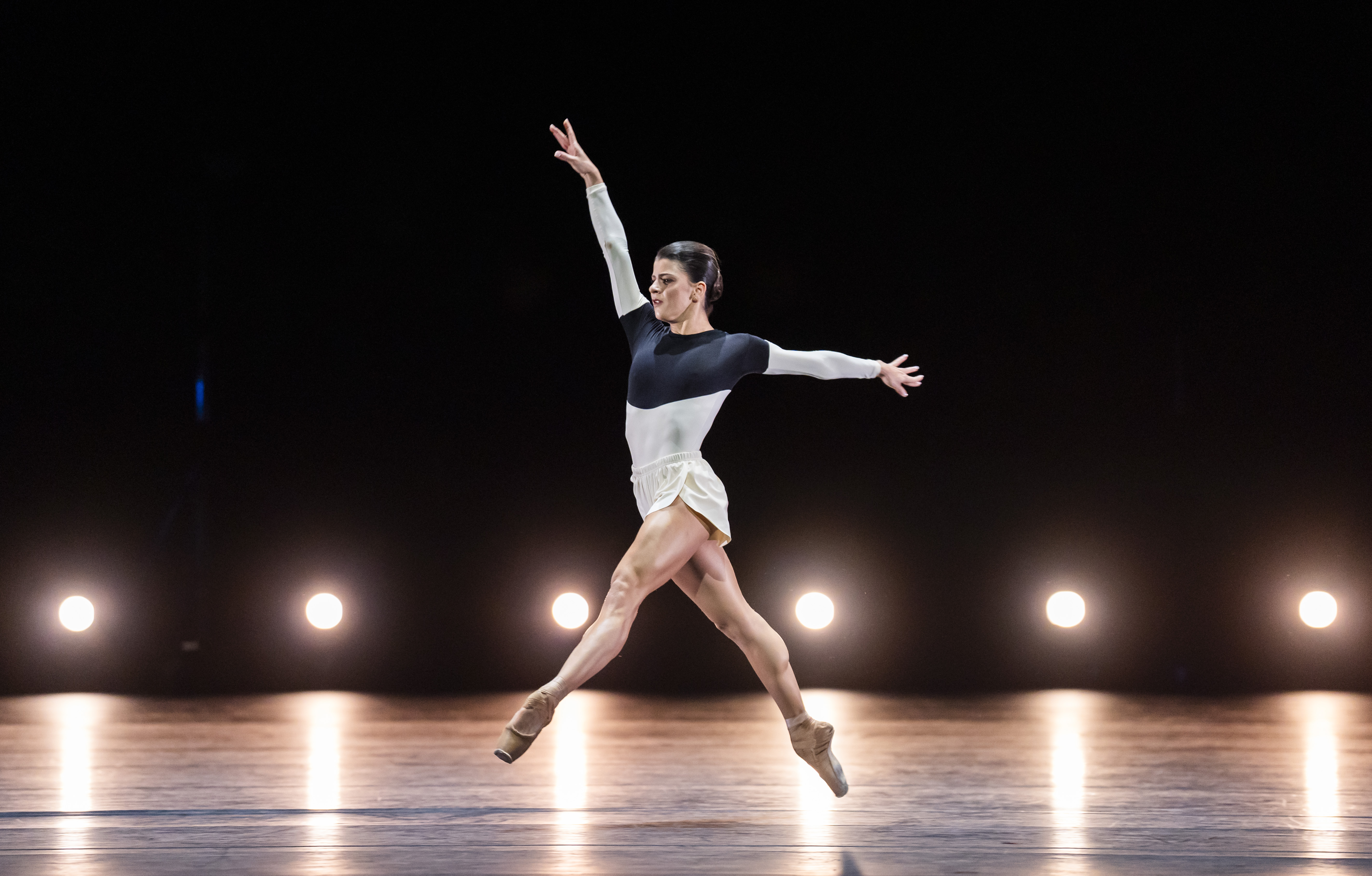 Mayara Magri performs on a dark stage. Behind her is a row of lights on the floor. She wears white shorts and a blue and white leotard. She is mid-jump with arms outstretched and one leg pointed forward, the other backwads. (2 of 5)