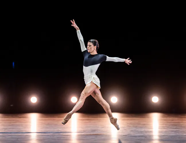 Mayara Magri performs on a dark stage. Behind her is a row of lights on the floor. She wears white shorts and a blue and white leotard. She is mid-jump with arms outstretched and one leg pointed forward, the other backwads. (2 of 5)