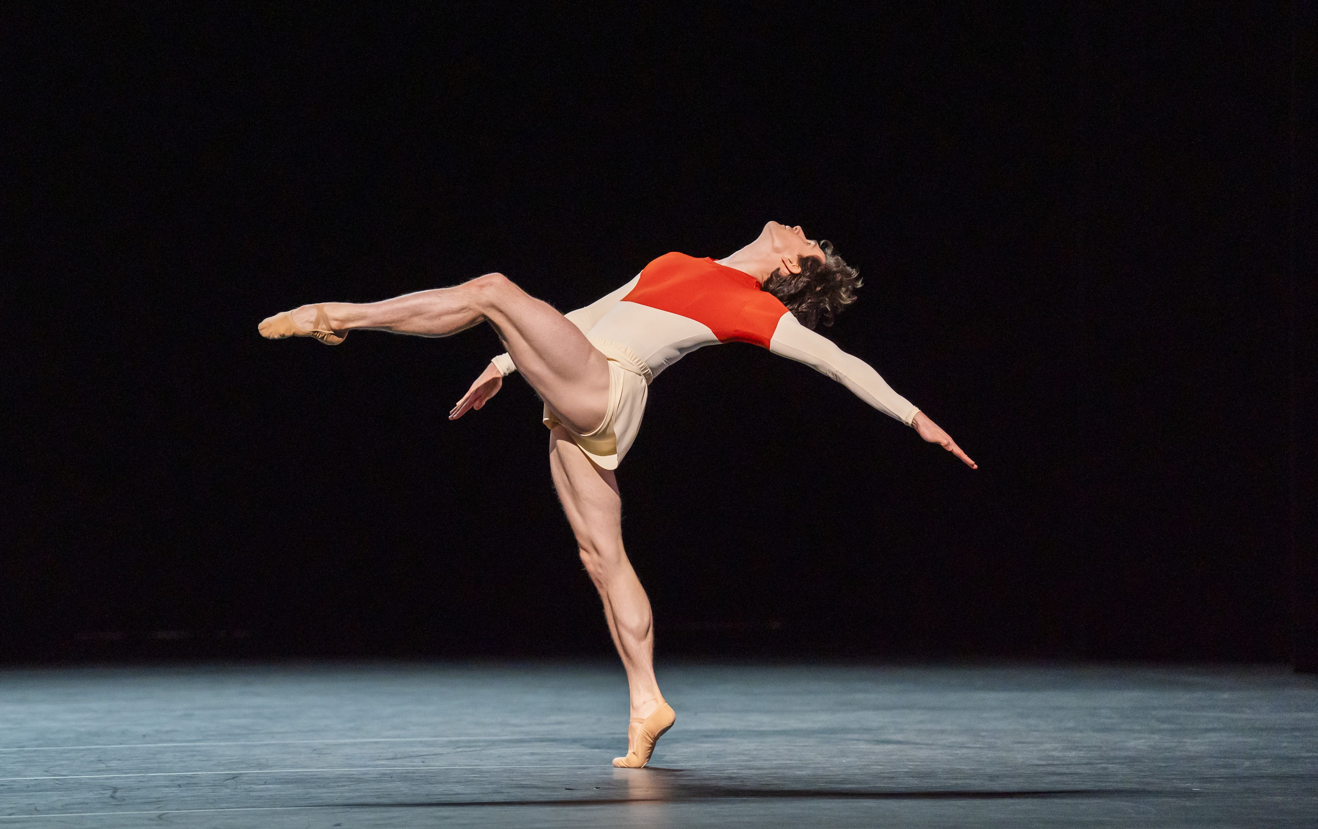 William Bracewell dances on stage. He wears white shorts and red and white leotard. He stands on demi-pointe with one leg outstretched before him. He casts one arm behind himself. (3 of 5)