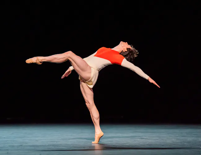 William Bracewell dances on stage. He wears white shorts and red and white leotard. He stands on demi-pointe with one leg outstretched before him. He casts one arm behind himself. (3 of 5)