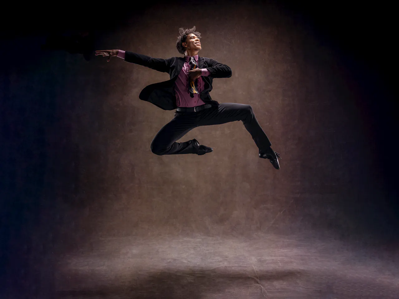 Carlos Acosta leaps midair in front of a dark brown backdrop. He wears a suit with a purple shirt. His right arm is outstretched behind him.