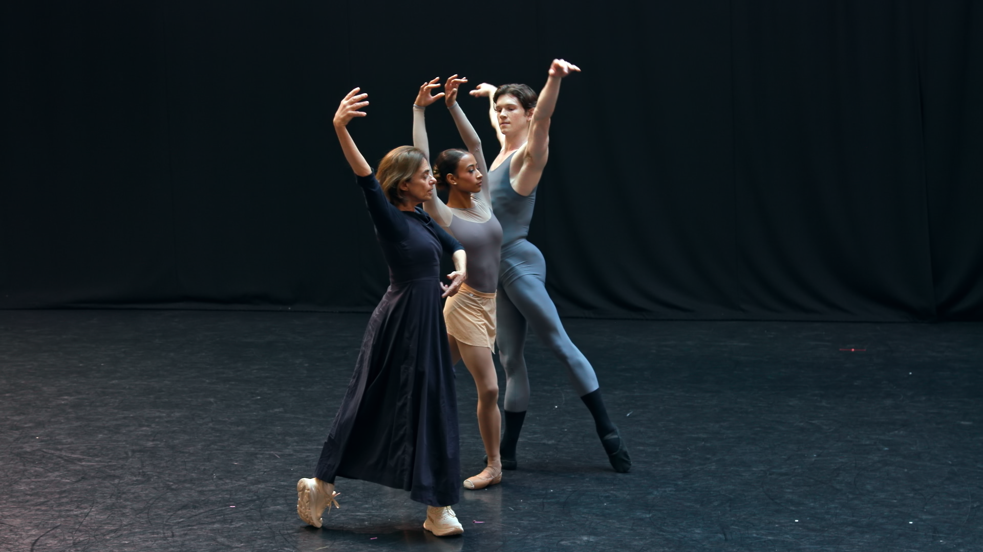 Three dancers stand in the middle of a dark stage. They stand near each other with their arms stretched above their heads and legs pointed out to the back. 