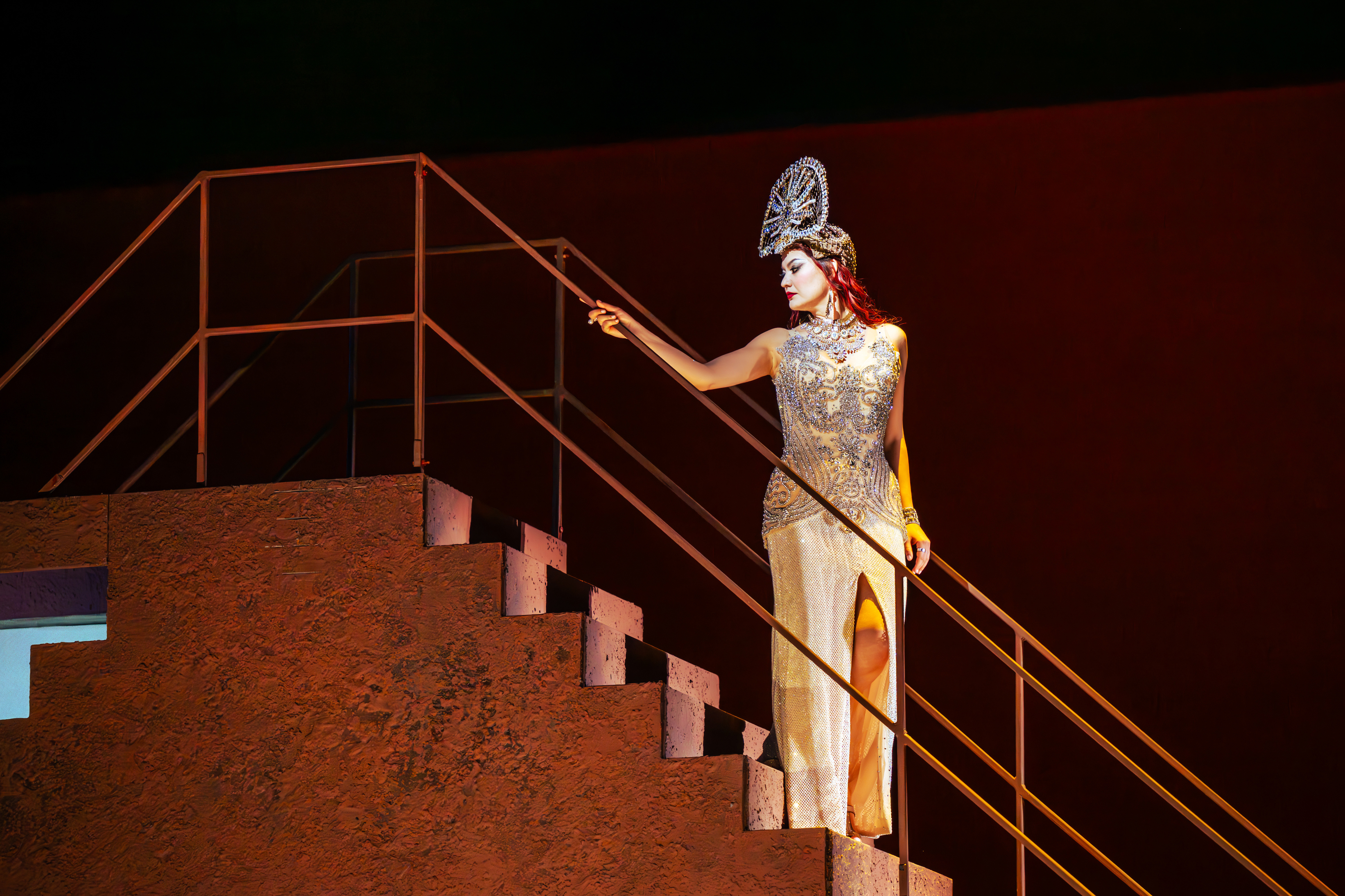 A woman in a glittering dress stands on a flight of rusty iron stairs. She is wearing a large, glittering headdress. (2 of 6)