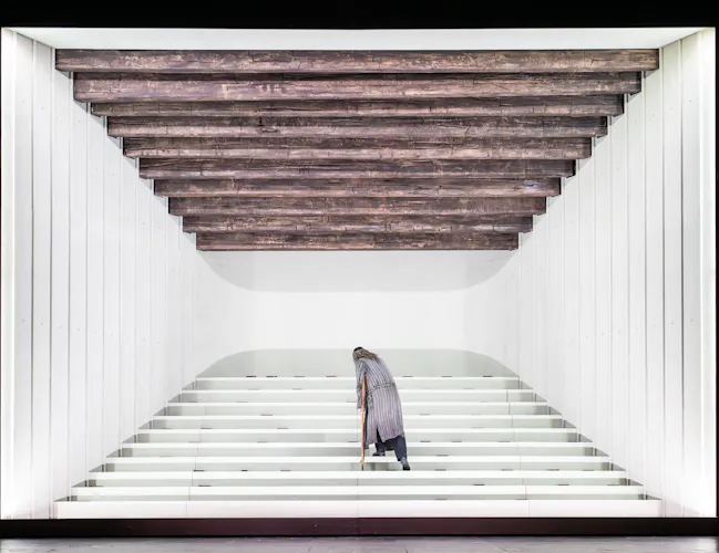 A man stands in a white box on a set of stairs. He has his back to the audience. (3 of 6)