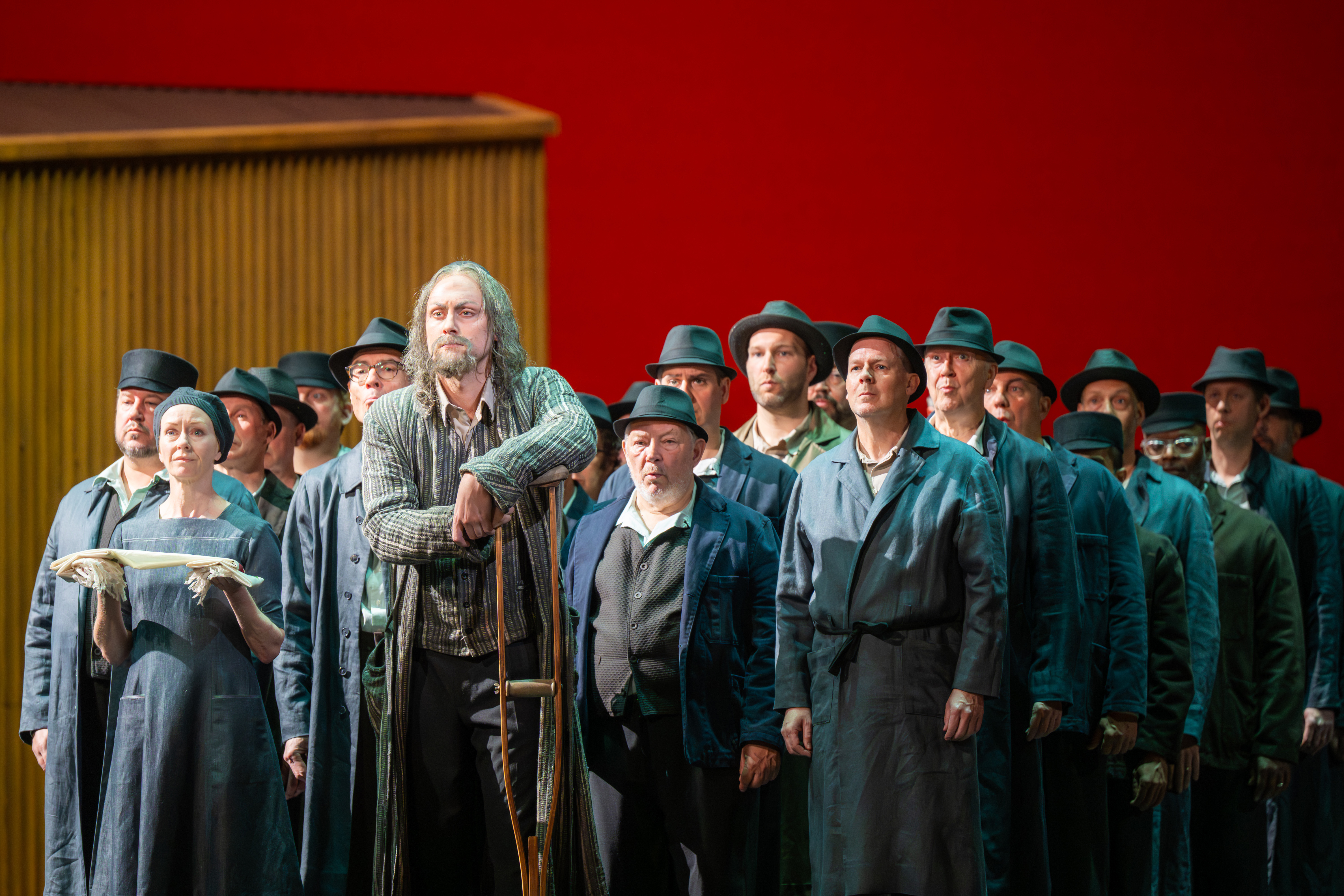 A group of performers stand on a stage with a red backdrop. At the front a man in a long grey coat leans on a crutch. Behind the people are wearing workers overalls and hats. (5 of 6)