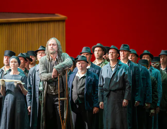 A group of performers stand on a stage with a red backdrop. At the front a man in a long grey coat leans on a crutch. Behind the people are wearing workers overalls and hats. (5 of 6)