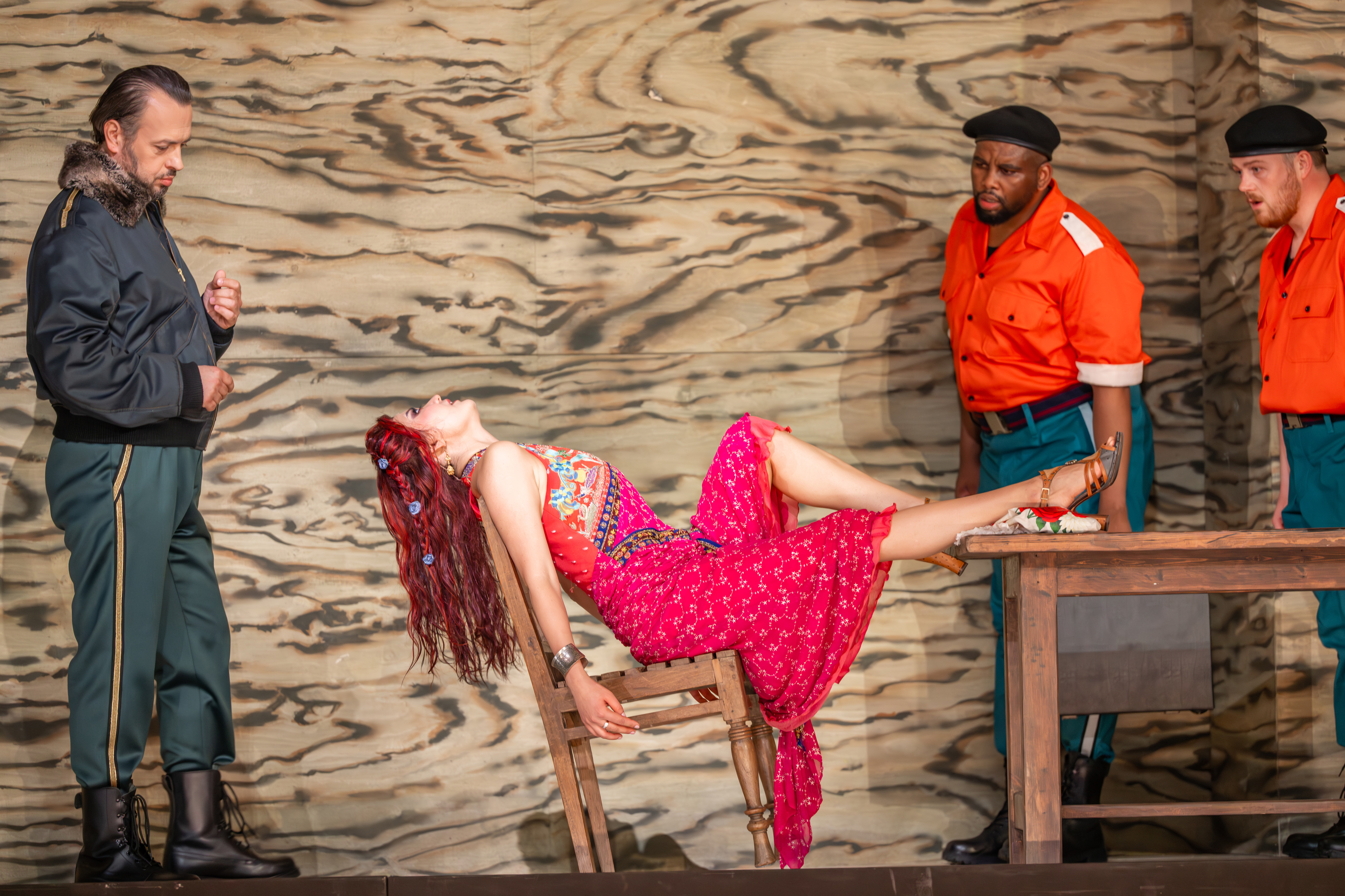 A woman in a pink dress leans back in her chair. Behind her is a man in army fatigues. Two guards stand over her. (6 of 6)