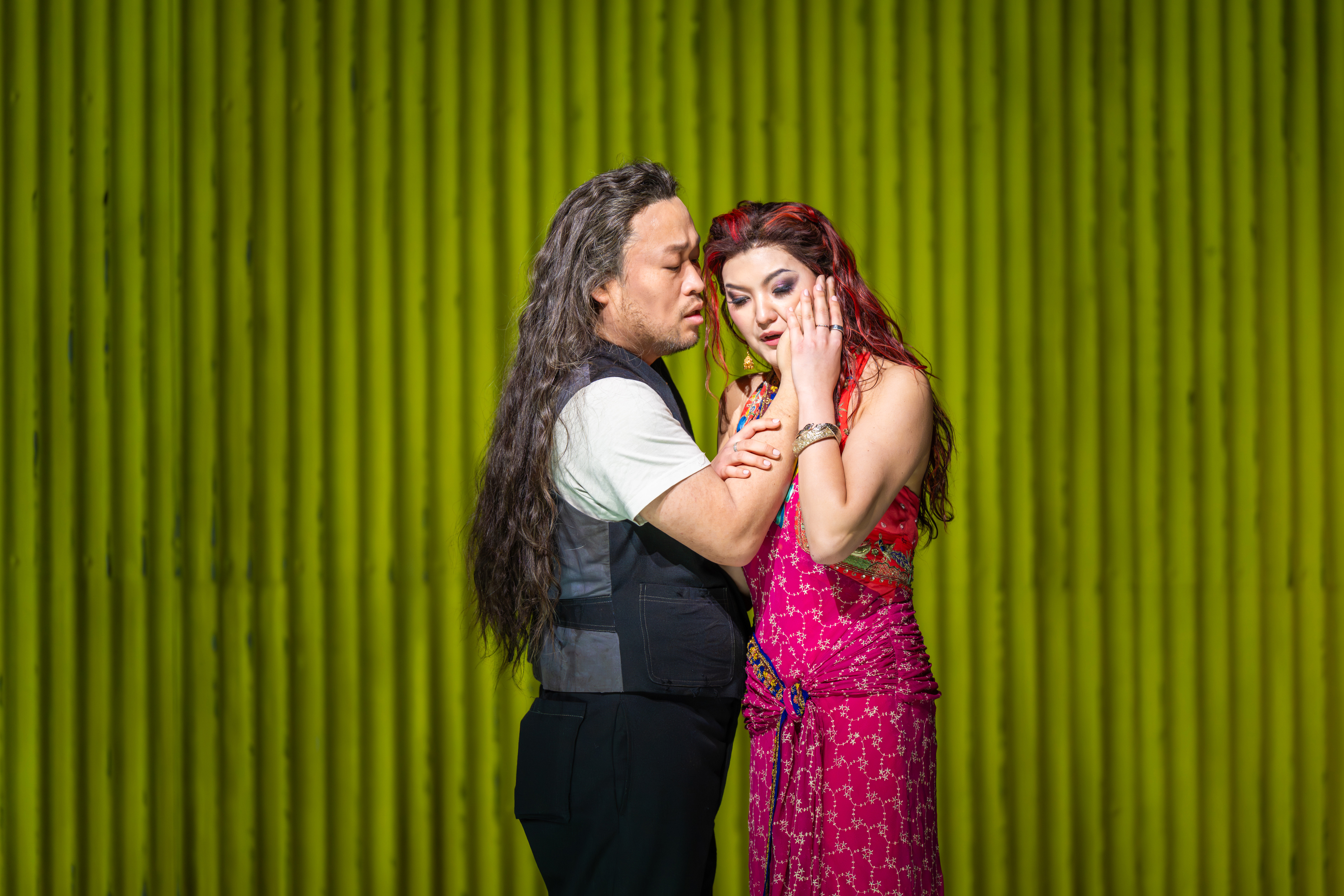 A man with long hair stands close to a woman in a pink dress. They are caressing each other's faces. They stand against a background of green corrugated iron. (4 of 6)