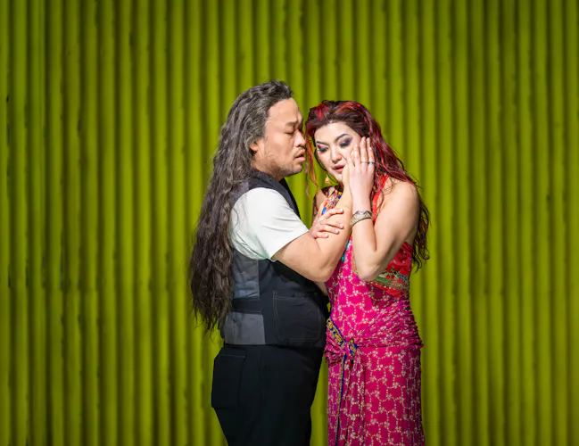 A man with long hair stands close to a woman in a pink dress. They are caressing each other's faces. They stand against a background of green corrugated iron. (4 of 6)