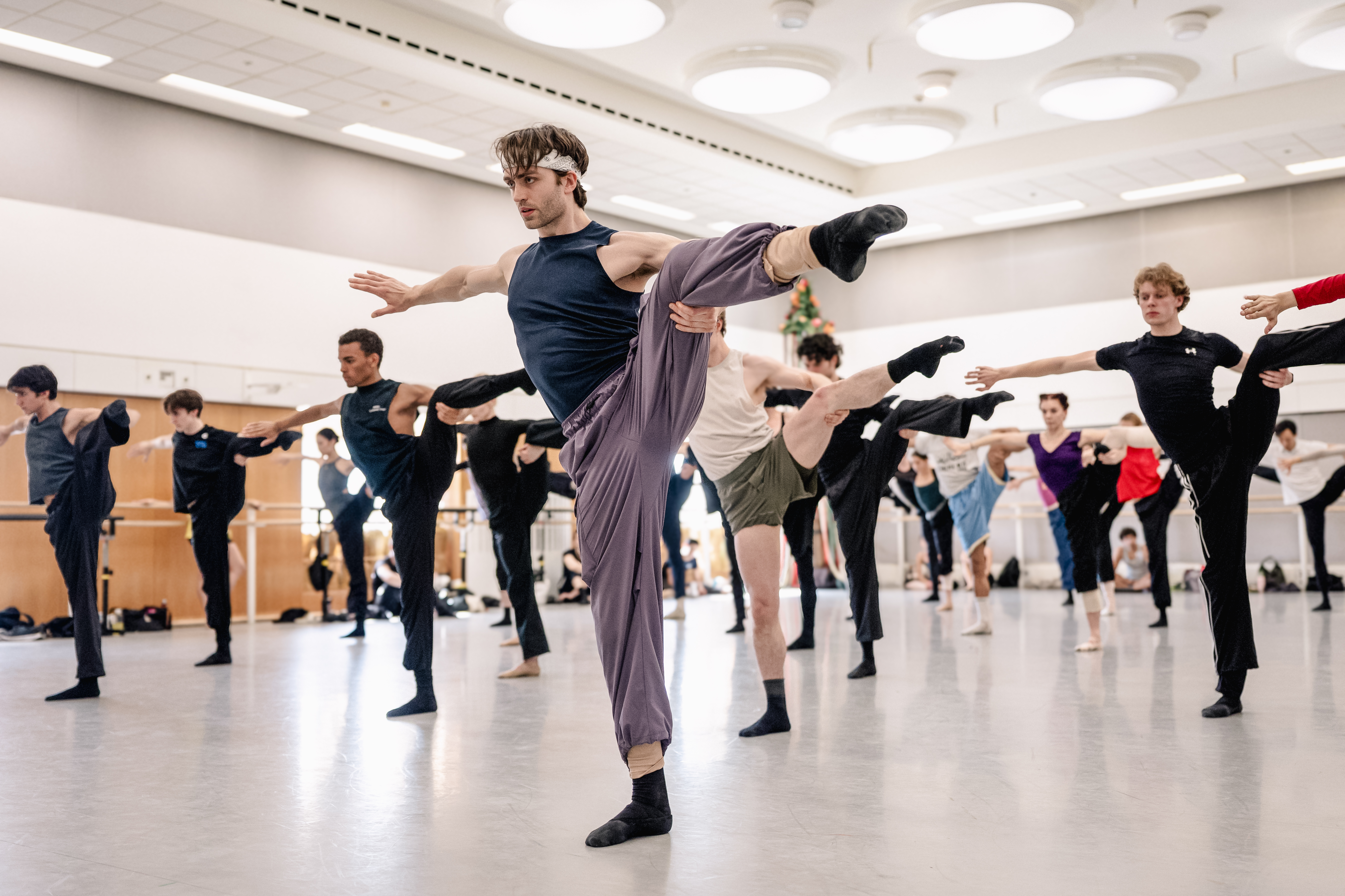 Matthew Ball in rehearsal for León and Lightfoot’s Salle de danse. He wears purple exercise trousers and stands on one leg with the other leg and his arms outstretched. Behind him other members of the Company practice.