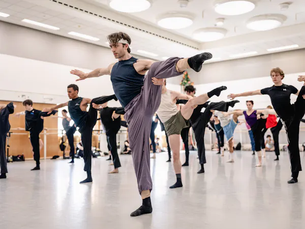 Matthew Ball in rehearsal for León and Lightfoot’s Salle de danse. He wears purple exercise trousers and stands on one leg with the other leg and his arms outstretched. Behind him other members of the Company practice.
