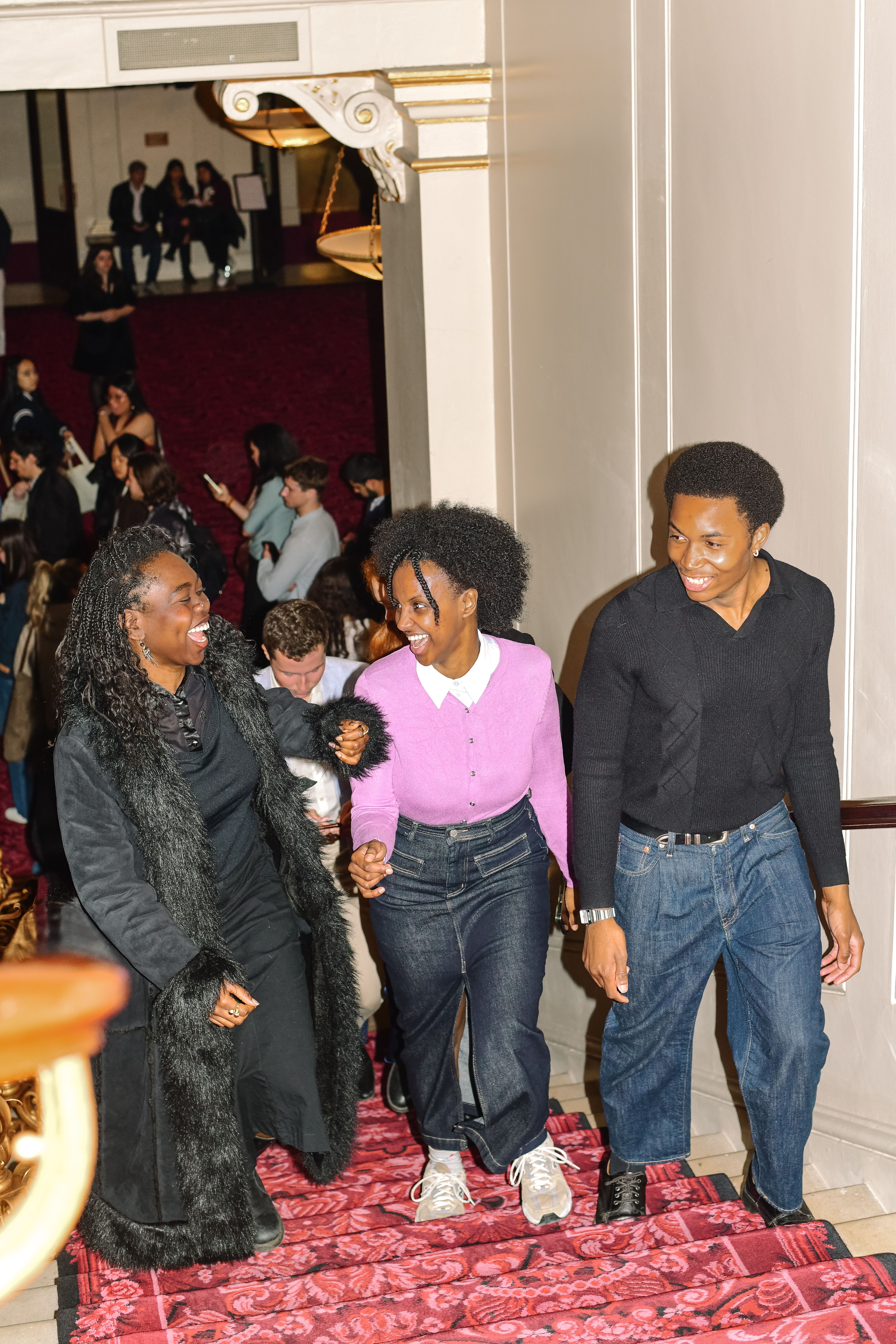 Three people walk up red decorative carpeted stairs in a theatre space. Behind them groups of people congregate at the bottom of the stairwell. (1 of 9)