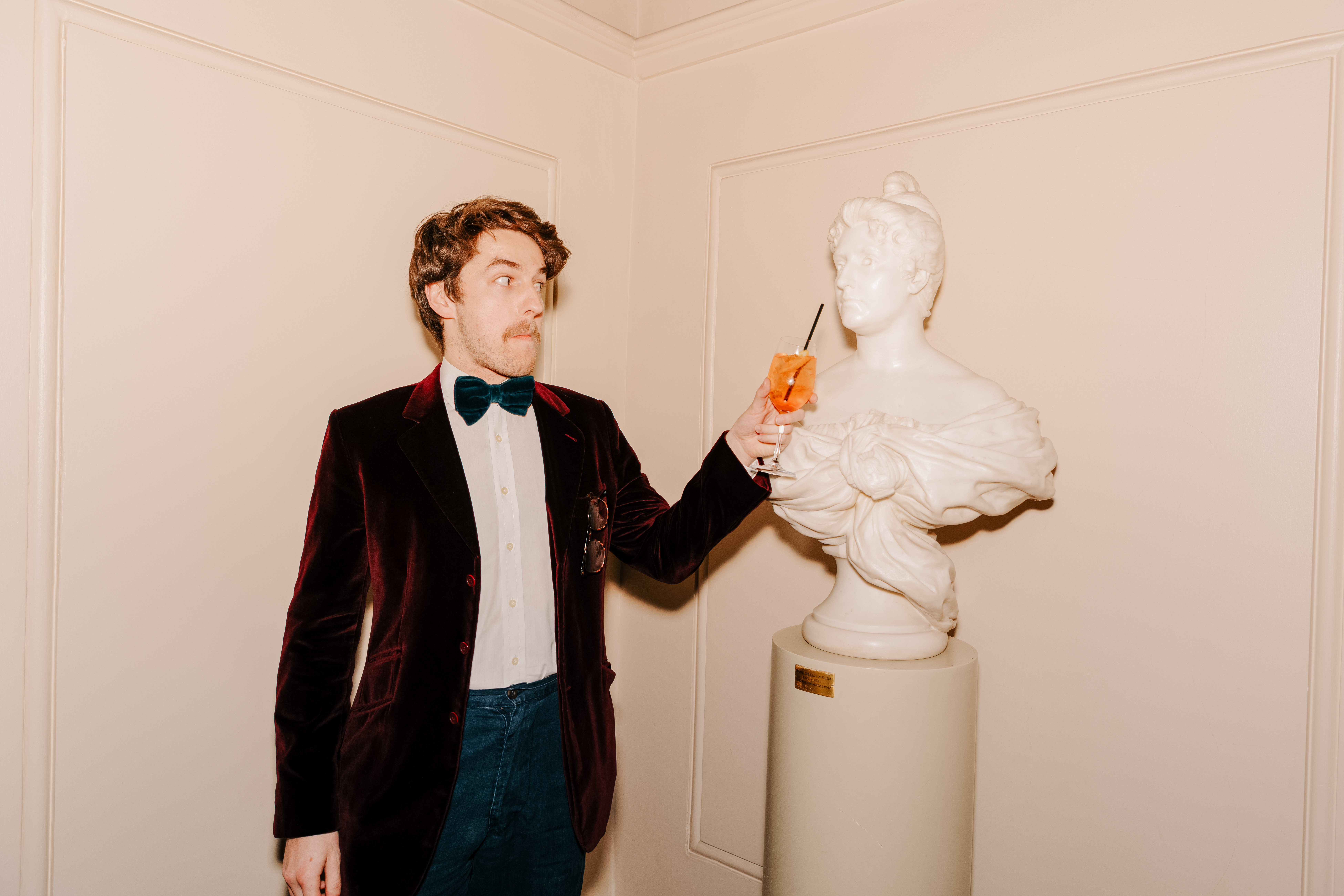 A person holds a glass of aperol with a straw in up to a bust sculpture as though they are drinking from it. (7 of 9)