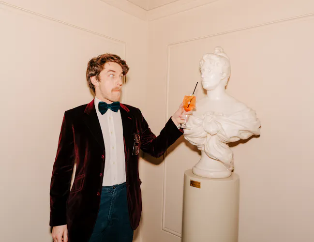 A person holds a glass of aperol with a straw in up to a bust sculpture as though they are drinking from it. (7 of 9)