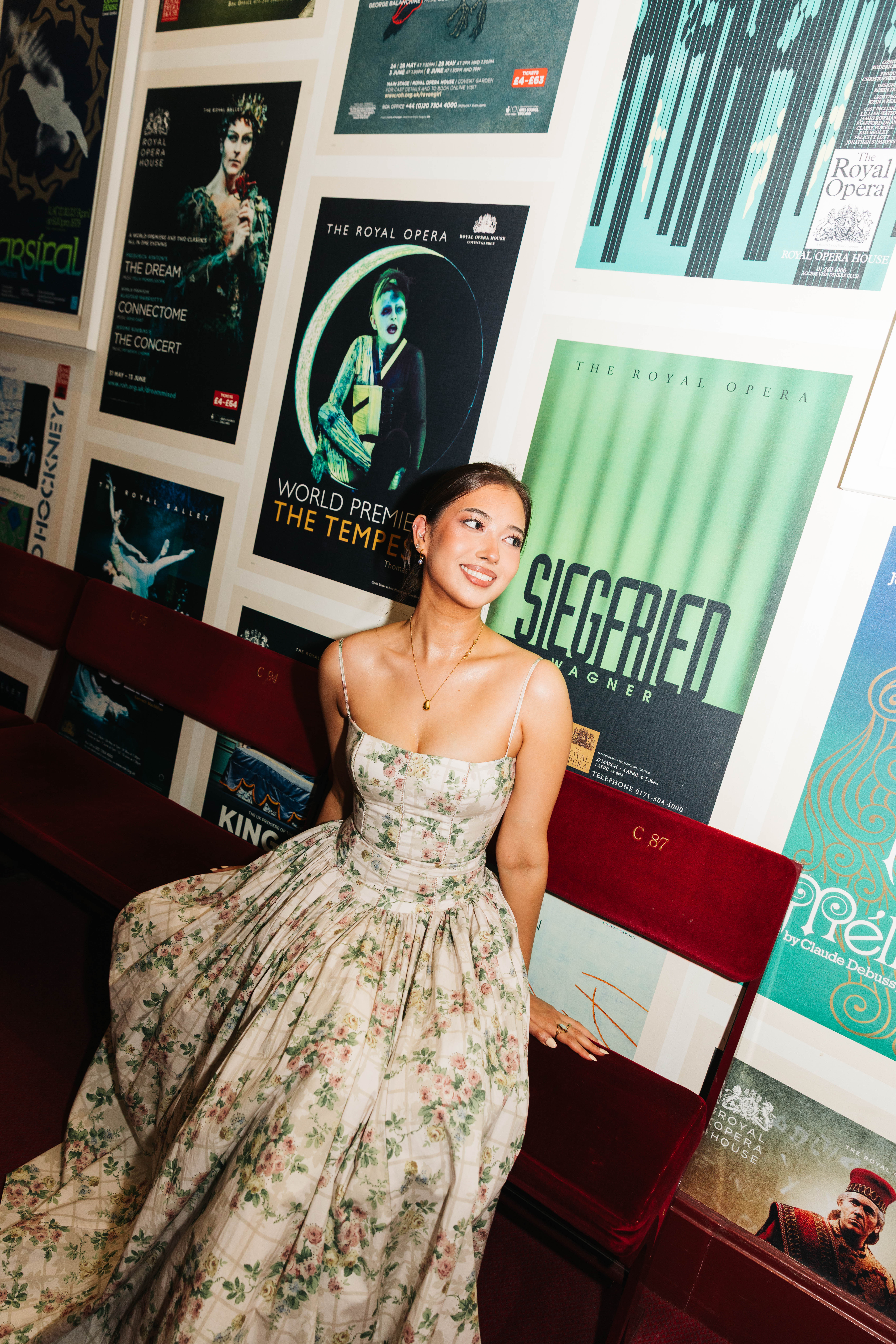 A person in a floral evening dress sits on red velvet theatre seats in front of a wall covered in vintage posters for operas at The Royal Opera House. (8 of 9)