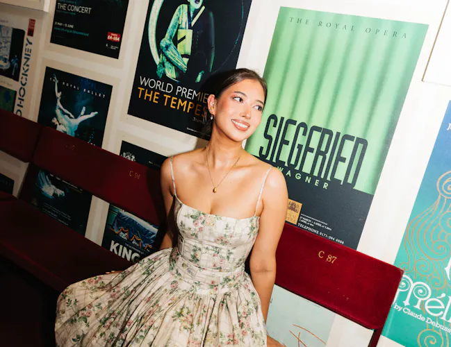 A person in a floral evening dress sits on red velvet theatre seats in front of a wall covered in vintage posters for operas at The Royal Opera House. (8 of 9)